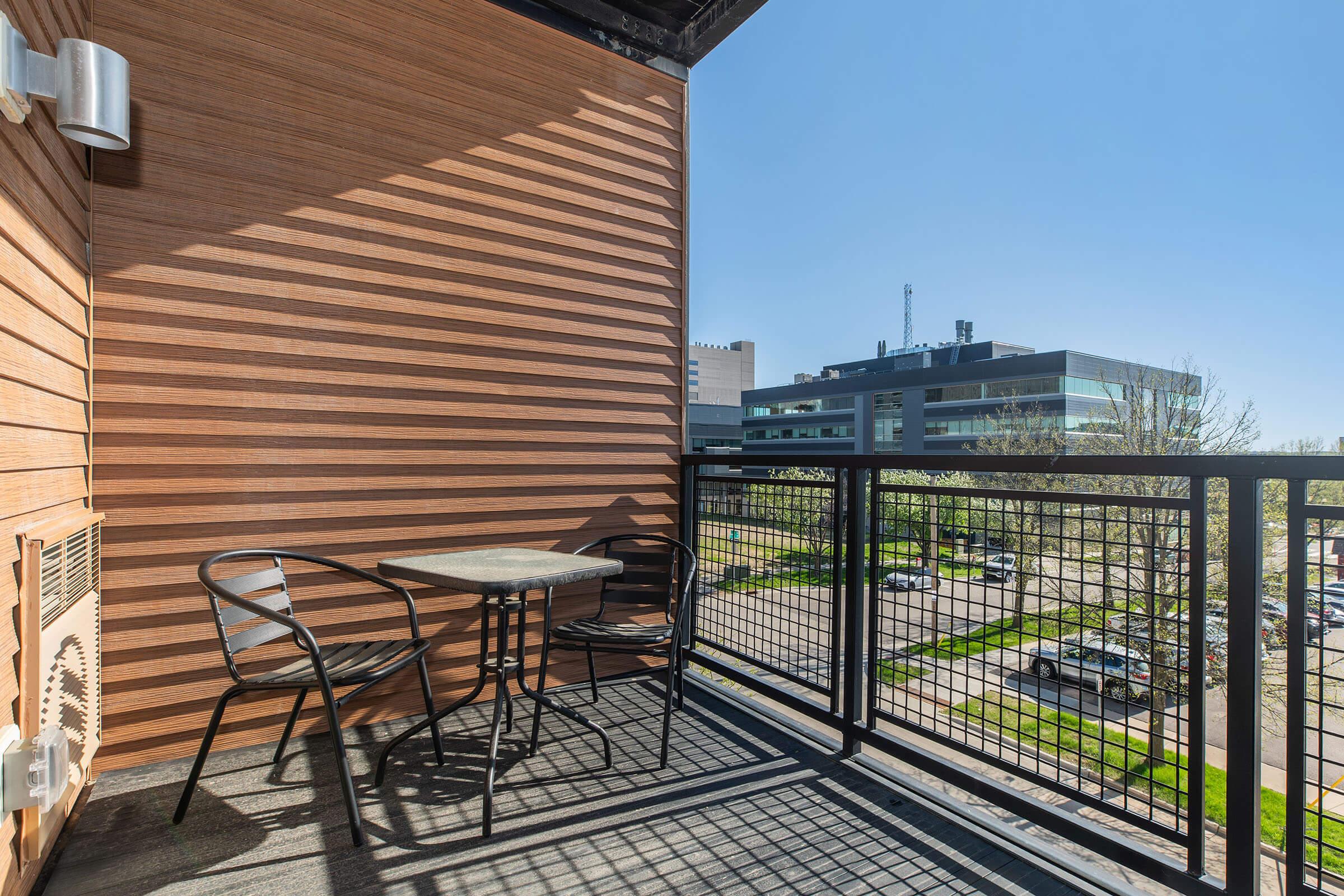 A small balcony featuring a round table and two chairs, with a clear blue sky in the background. The balcony has a black railing and wooden paneling, overlooking a parking lot and nearby buildings. The scene is bright and inviting, suggesting a cozy outdoor space for relaxation.