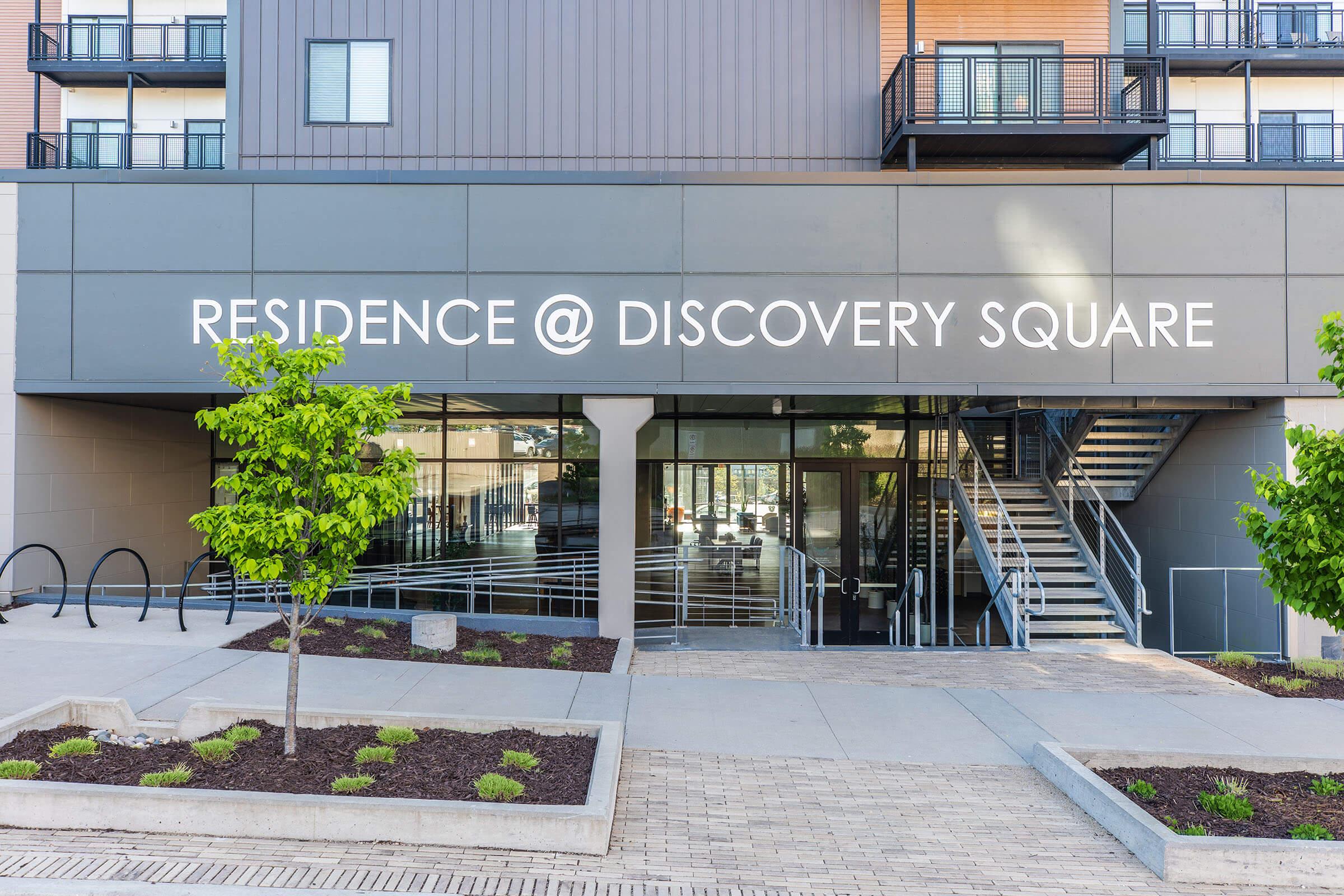 Facade of an apartment building with the sign "RESIDENCE @ DISCOVERY SQUARE" prominently displayed. The entrance features large glass doors and surrounding landscaped areas with small trees, while stairs lead to an upper level. The overall design is modern and welcoming.