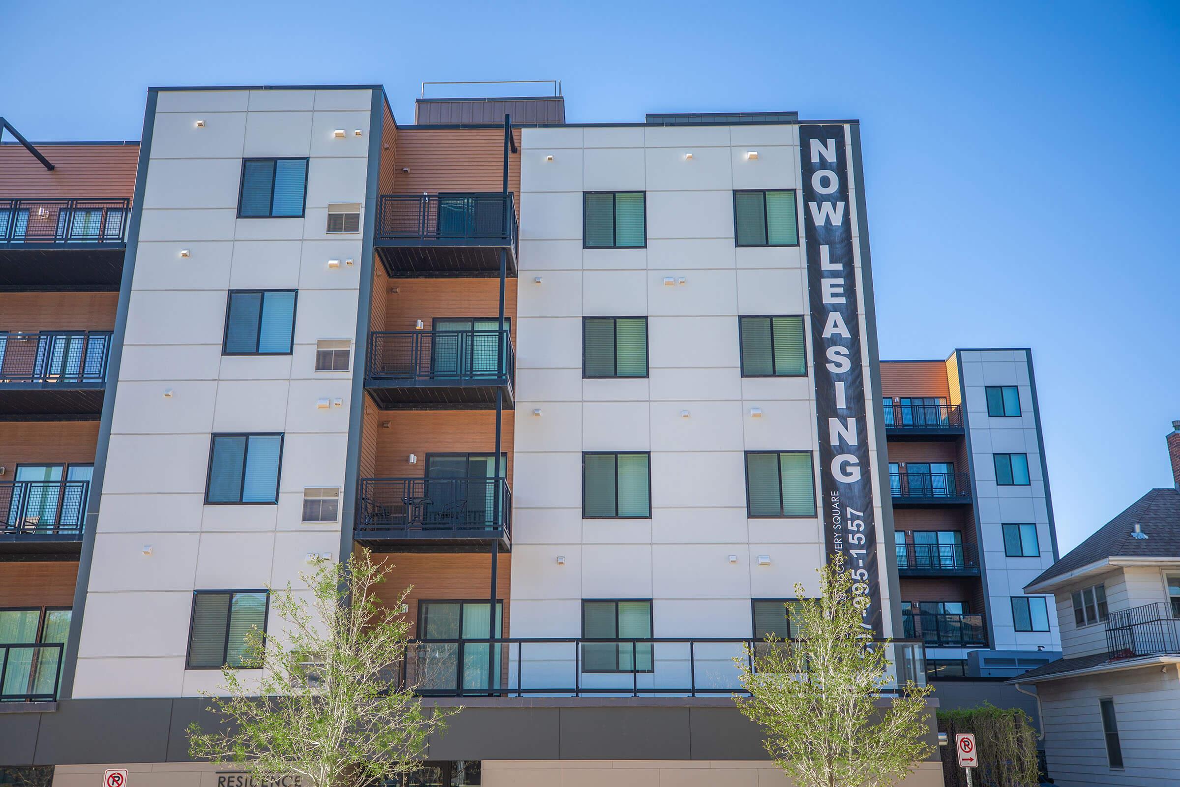 A modern apartment building featuring multiple floors with large windows and a "NOW LEASING" banner prominently displayed on the side. The exterior combines white and wooden elements. Small trees are visible at the base of the building, and the sky is clear and blue.