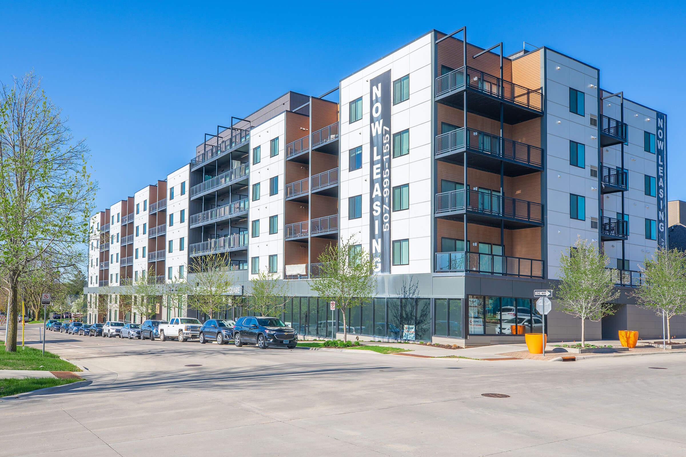 Modern apartment building with multiple stories featuring balconies, large windows, and a "NOW LEASING" sign. The structure has a mix of white and dark-colored panels, surrounded by young trees and parked cars along the street on a clear blue sky day.