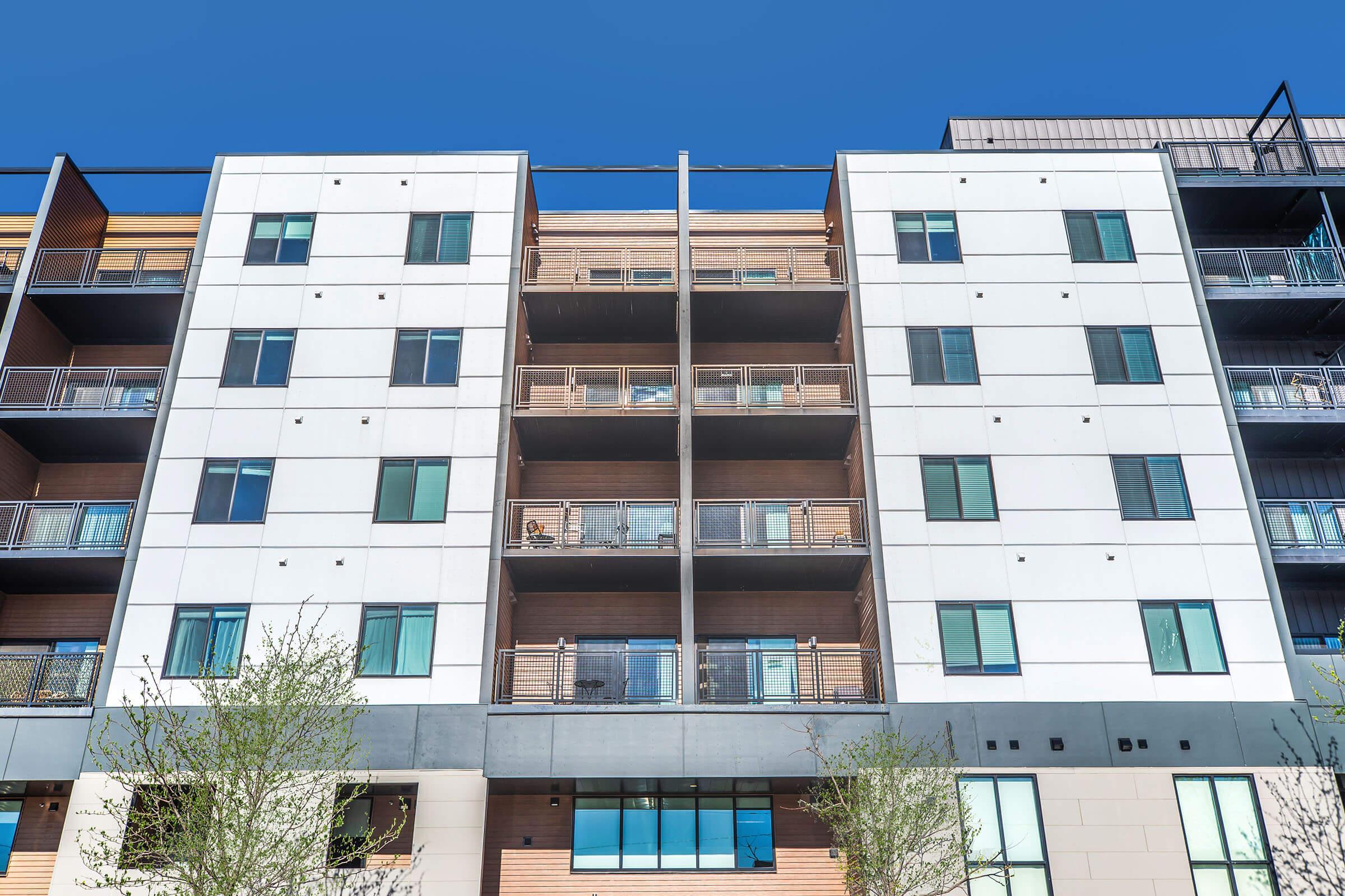 Modern multi-story apartment building featuring a combination of white and wooden panel exteriors, with multiple balconies. The upper floors are visible against a clear blue sky, and trees are in the foreground, adding greenery to the urban environment.