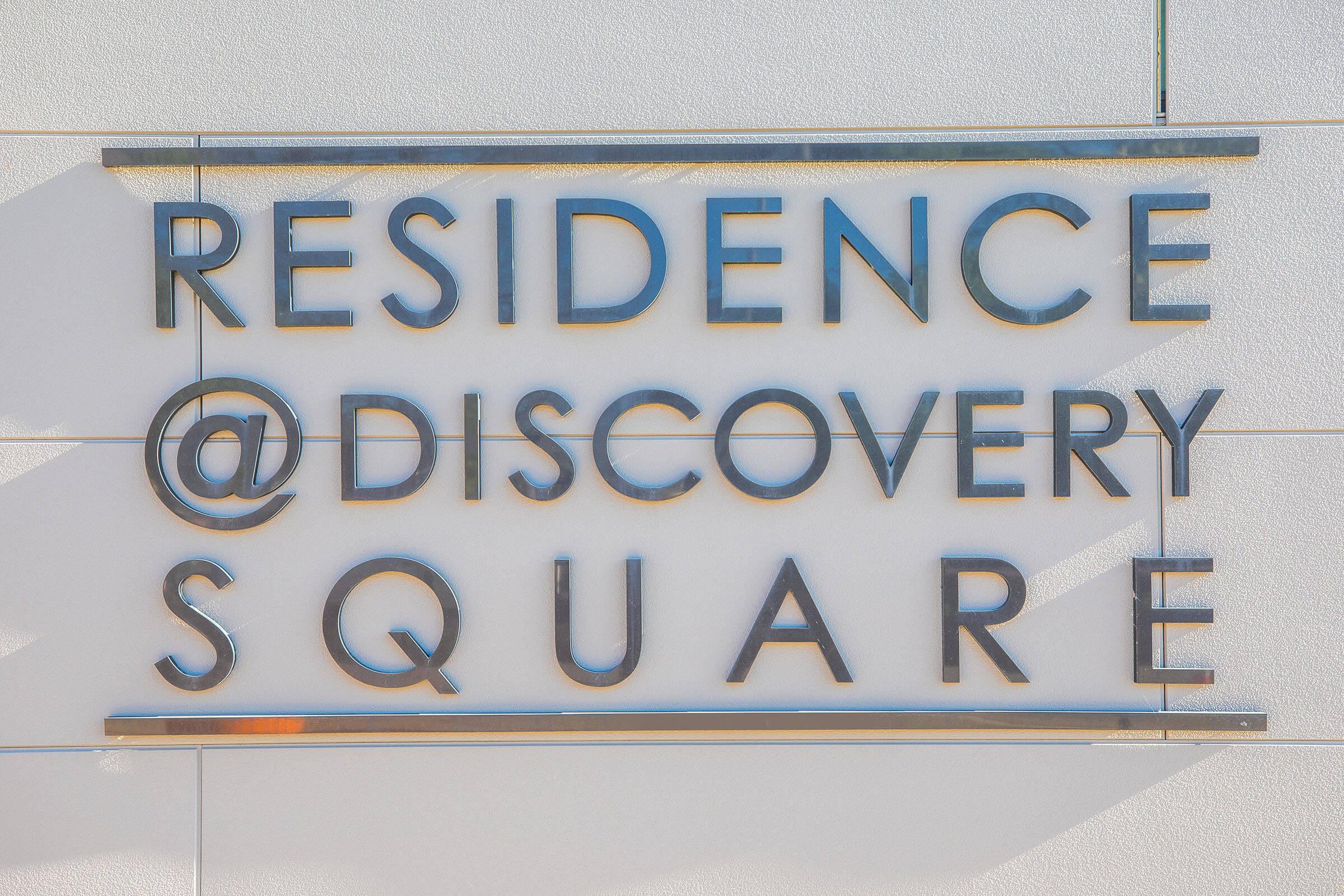 A sign displaying the text "RESIDENCE @ DISCOVERY SQUARE" mounted on a white wall. The letters are made of a metallic material that reflects light, creating shadows and highlighting the modern design of the sign.