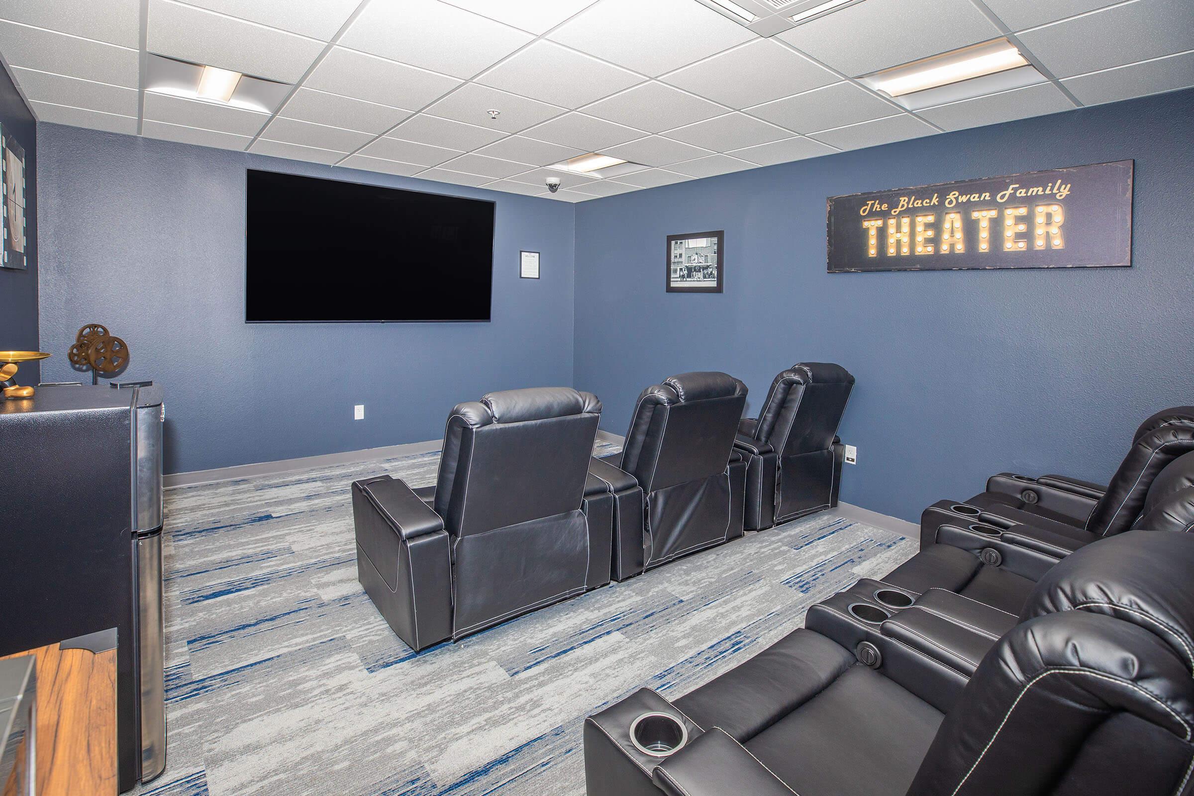 A cozy home theater room featuring dark blue walls, plush black recliner chairs, and a large flat-screen TV. A decorative sign reading "The Black Swan Family Theater" adds charm to the space. The floor is covered with stylish gray and blue carpet tiles, creating an inviting atmosphere for movie nights.