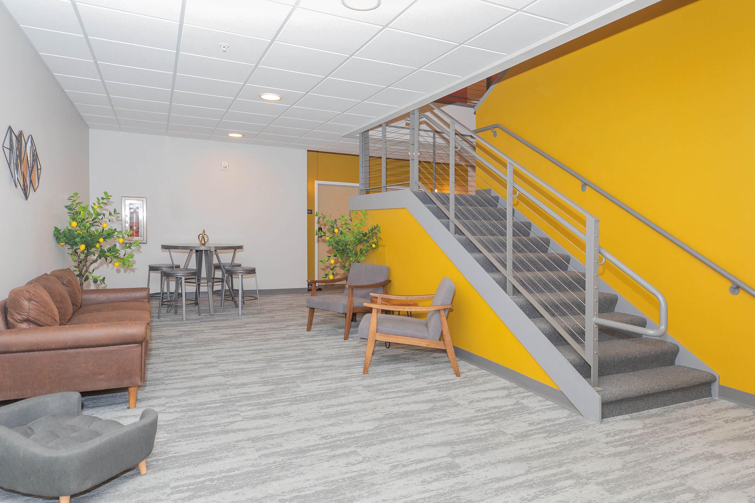 A modern interior space featuring a staircase against a vibrant yellow wall. The room includes a brown leather sofa, a wooden chair, and a small table with four chairs. Potted plants add greenery to the scene, and the flooring is light gray with a textured appearance.