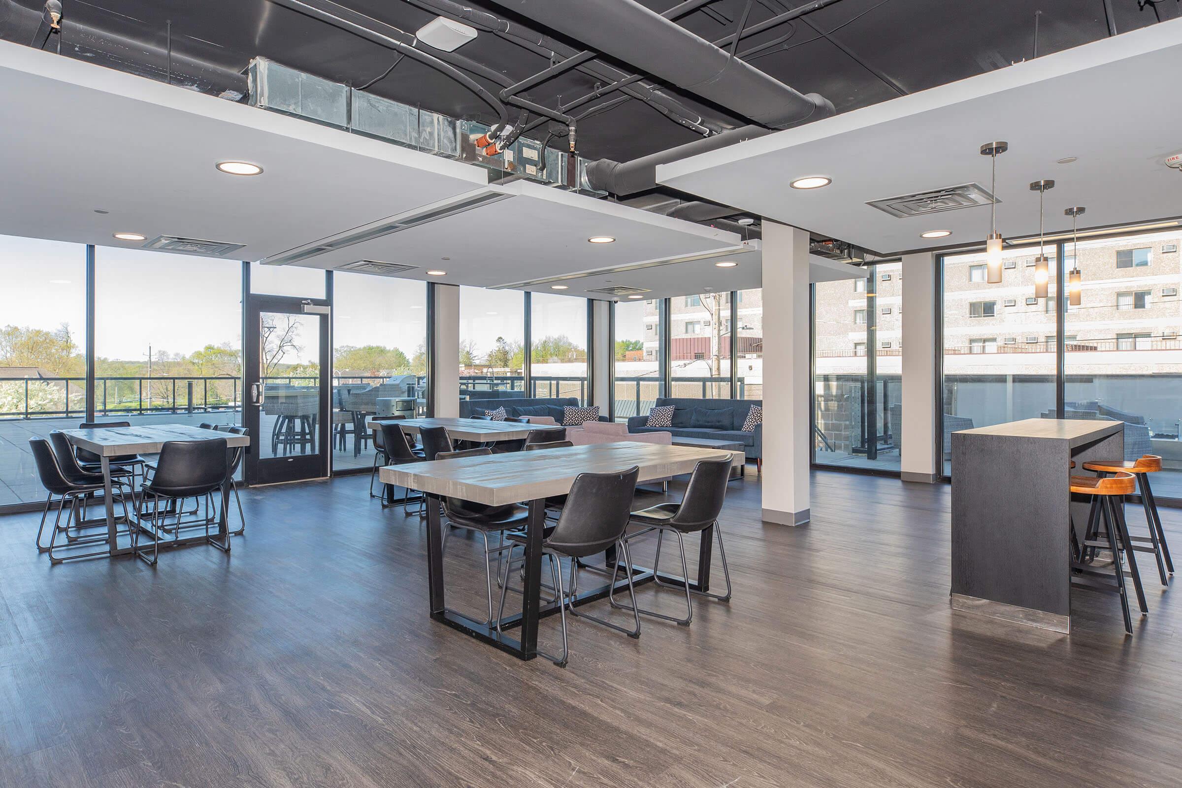 Modern, spacious lounge area with large windows letting in natural light. Features several tables and chairs arranged for dining or meetings, along with a small bar counter. The decor has a contemporary style, and there's an outdoor view of a patio or balcony area.