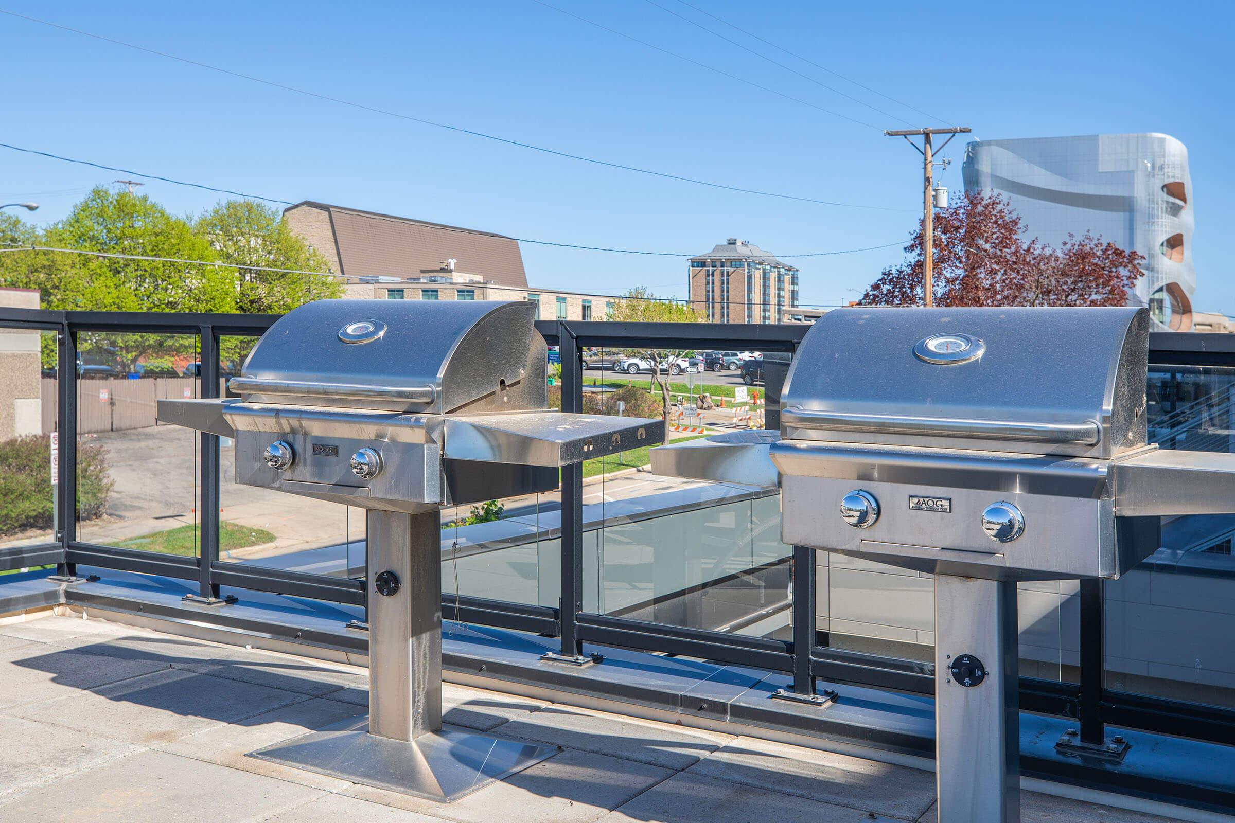 Two stainless steel grills positioned on a rooftop terrace, with a clear blue sky in the background. A modern building is partially visible behind the grills, along with trees and a cityscape in the distance. The scene suggests an outdoor cooking area for social gatherings.