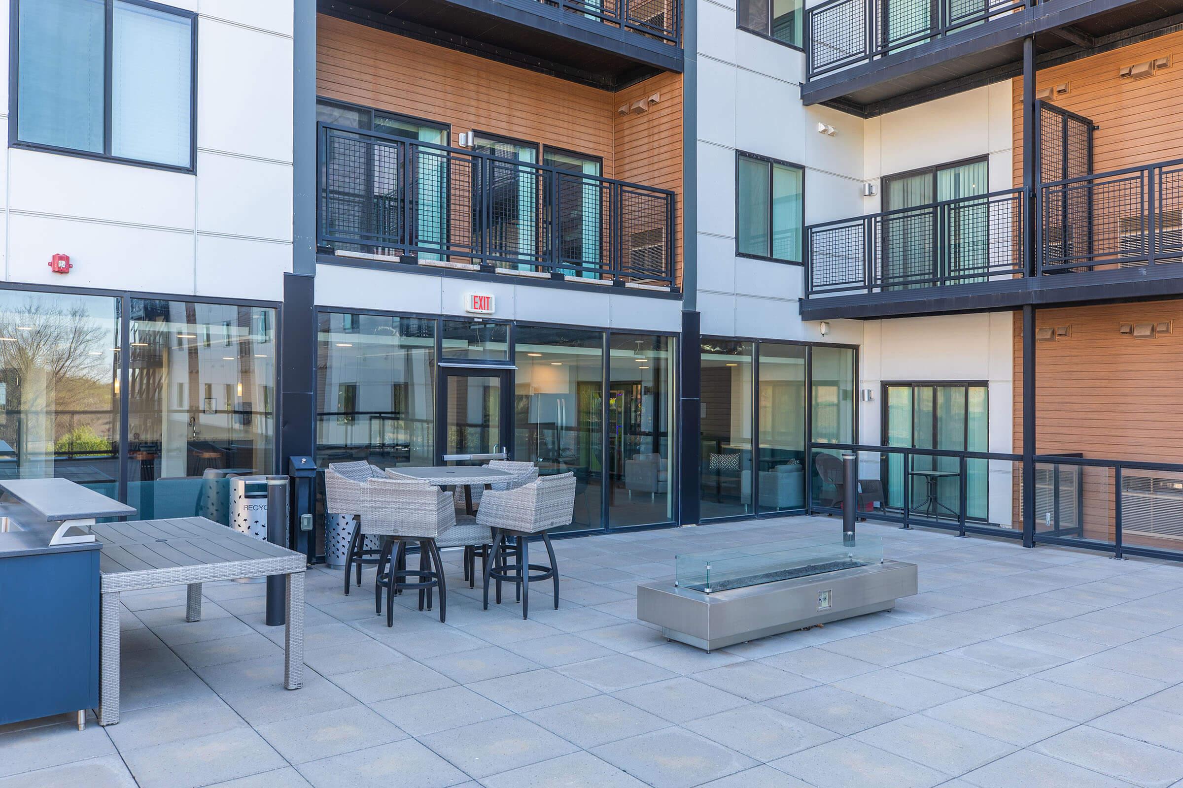 A modern outdoor patio area featuring sleek furniture, including a table with chairs, a fire pit, and a barbecue station. The space is surrounded by large glass windows of an apartment building, showcasing a contemporary design with a mix of materials. Natural light enhances the inviting atmosphere.