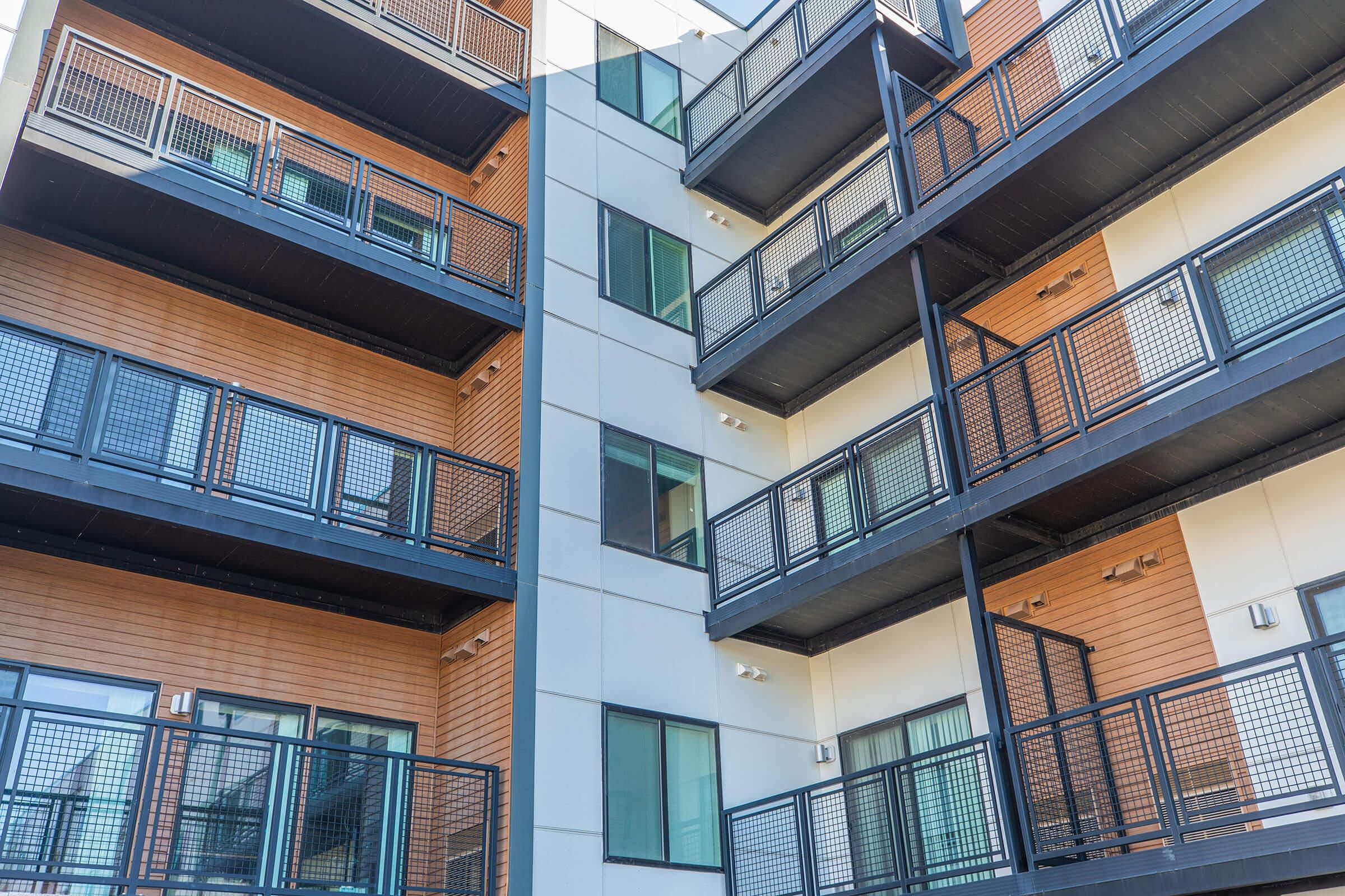 A modern apartment building featuring multiple stacked floors with balconies. The structure has a mix of wood and white exterior, large windows on each level, and metal railings. The design showcases a contemporary architectural style with a focus on light and openness.