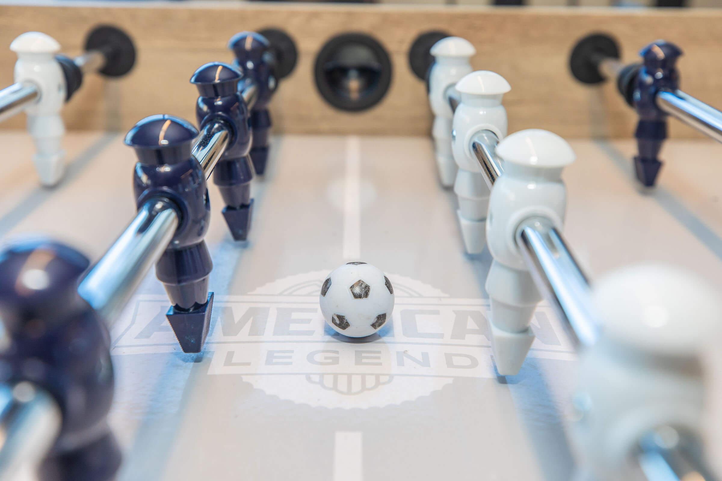 A close-up view of a foosball table featuring a small soccer ball positioned in the center, surrounded by blue and white player figures. The table surface has the text "American Legend" visible, along with the lines marking the playing area.