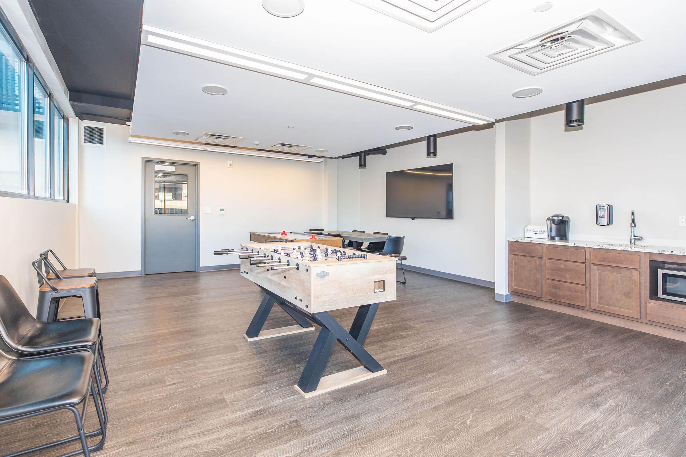 A modern recreational room featuring a foosball table centrally placed on dark wood flooring. There are black chairs along the sides, a kitchenette with cabinets, a countertop, and a coffee maker. A large flat-screen TV is mounted on the wall, and large windows provide natural light in the space.