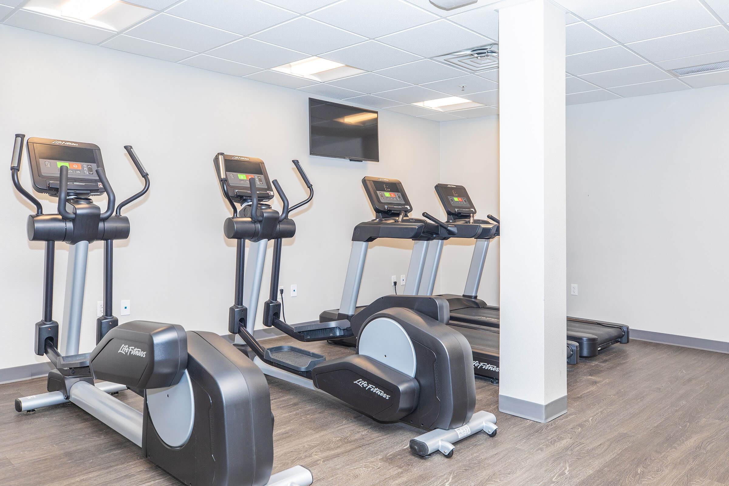 A fitness room featuring several modern elliptical machines and a treadmill. The space is well-lit with bright overhead lighting and has neutral-colored walls. A wall-mounted TV is visible, and the flooring is dark wood.