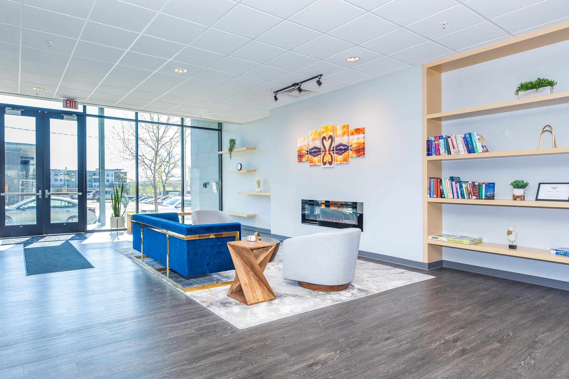 A modern lounge area featuring a blue sofa, a gray armchair, and a wooden coffee table. There’s an electric fireplace on the wall, surrounded by a small bookshelf filled with books. Large windows allow natural light to fill the space, showcasing outdoor views.