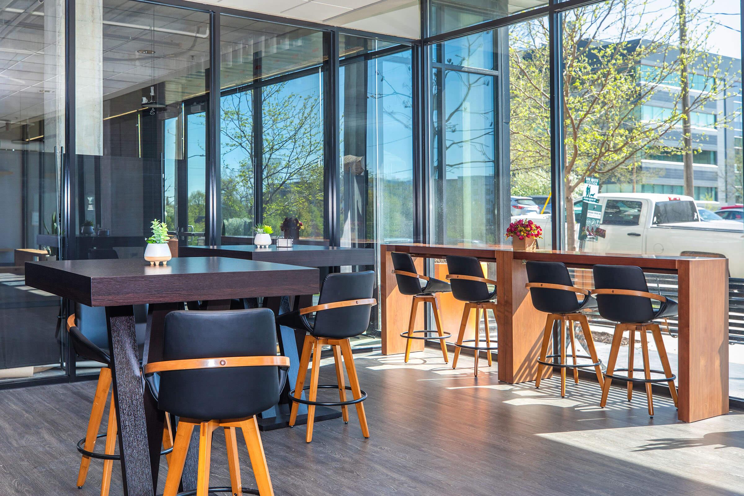 Modern indoor space featuring large windows with natural light, two types of seating arrangements: a tall wooden table with black and wooden bar stools, and a lower wooden table. Potted plants and a small flower arrangement add decor, with an outdoor view visible through the glass walls.