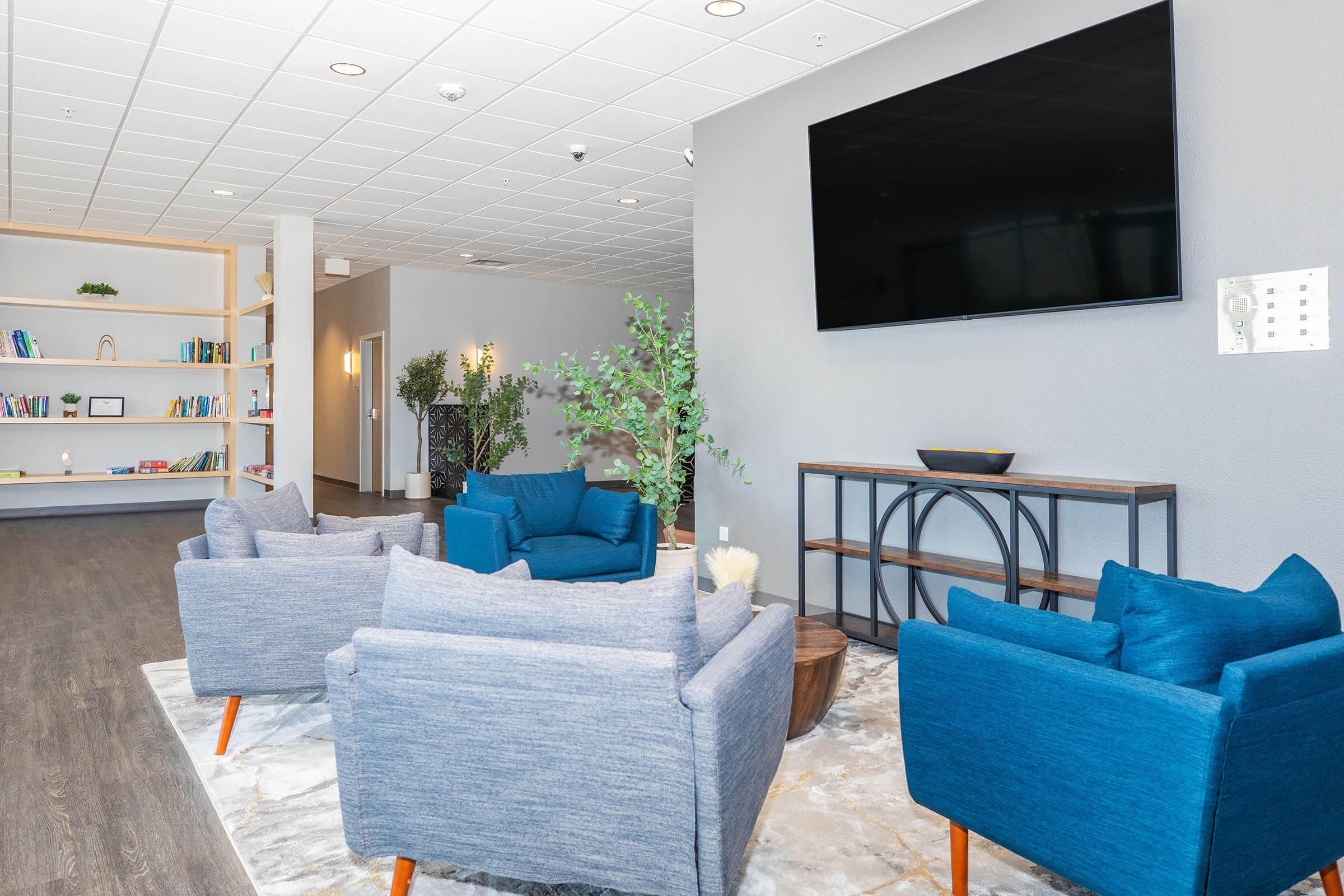 A modern lounge area featuring two blue armchairs and a round wooden coffee table on a marble floor. In the background, there is a mounted flat-screen TV and a bookshelf filled with books. The walls are a neutral gray, and there are plants and decor elements enhancing the inviting atmosphere.