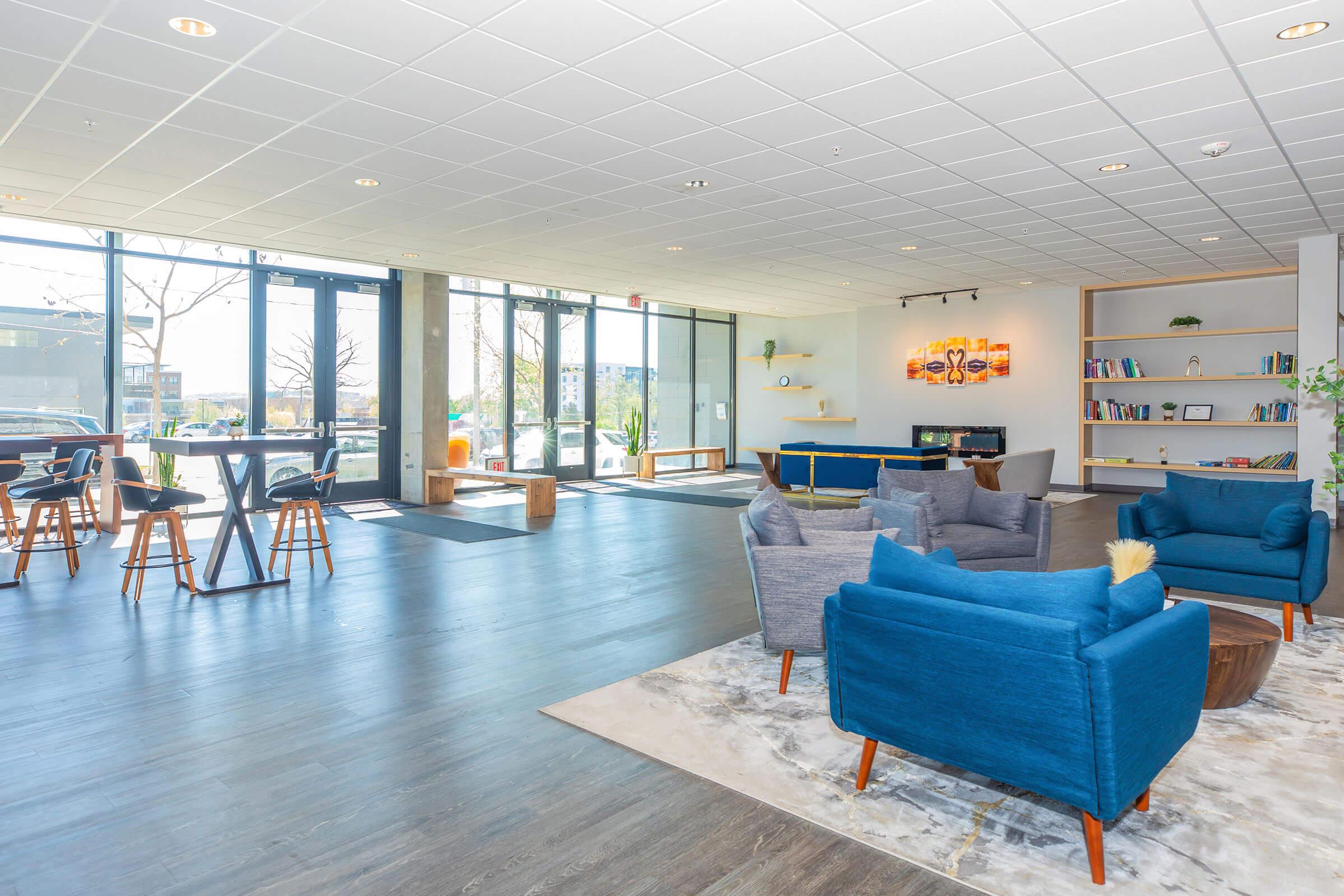 A modern, spacious lounge area featuring stylish blue armchairs, a coffee table, and a bookshelf. Large windows allow natural light, and there are decorative elements like wall art and plants. Tables and chairs are arranged for socializing, creating a welcoming atmosphere.