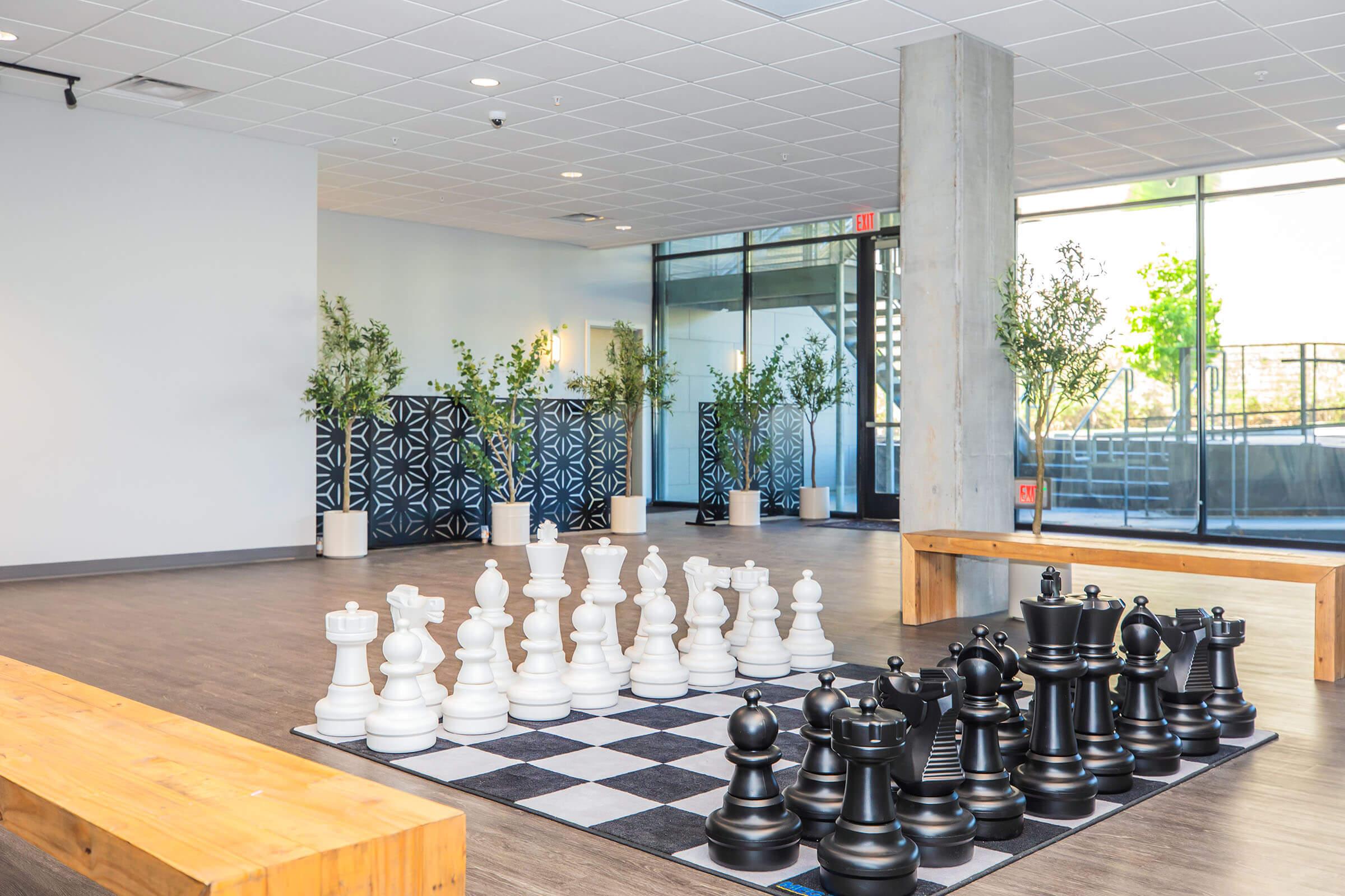 A large indoor space featuring a chessboard with oversized black and white chess pieces. The area is brightly lit, with large windows, greenery in the background, and wooden benches lining the space. The modern design emphasizes a clean and open atmosphere.