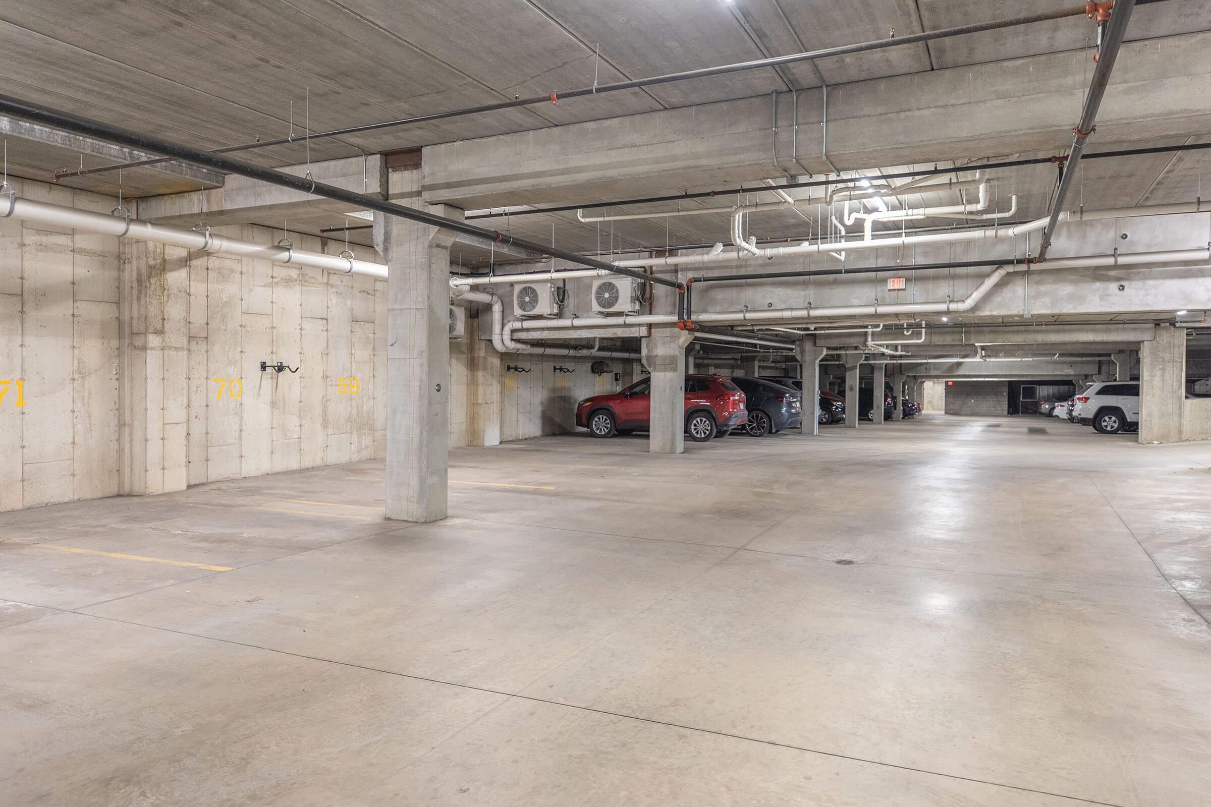 An empty underground parking garage featuring concrete walls and floors. Various parking spaces are marked, with several cars parked in designated areas. The garage is well-lit, with exposed pipes and ventilation systems visible on the ceiling.