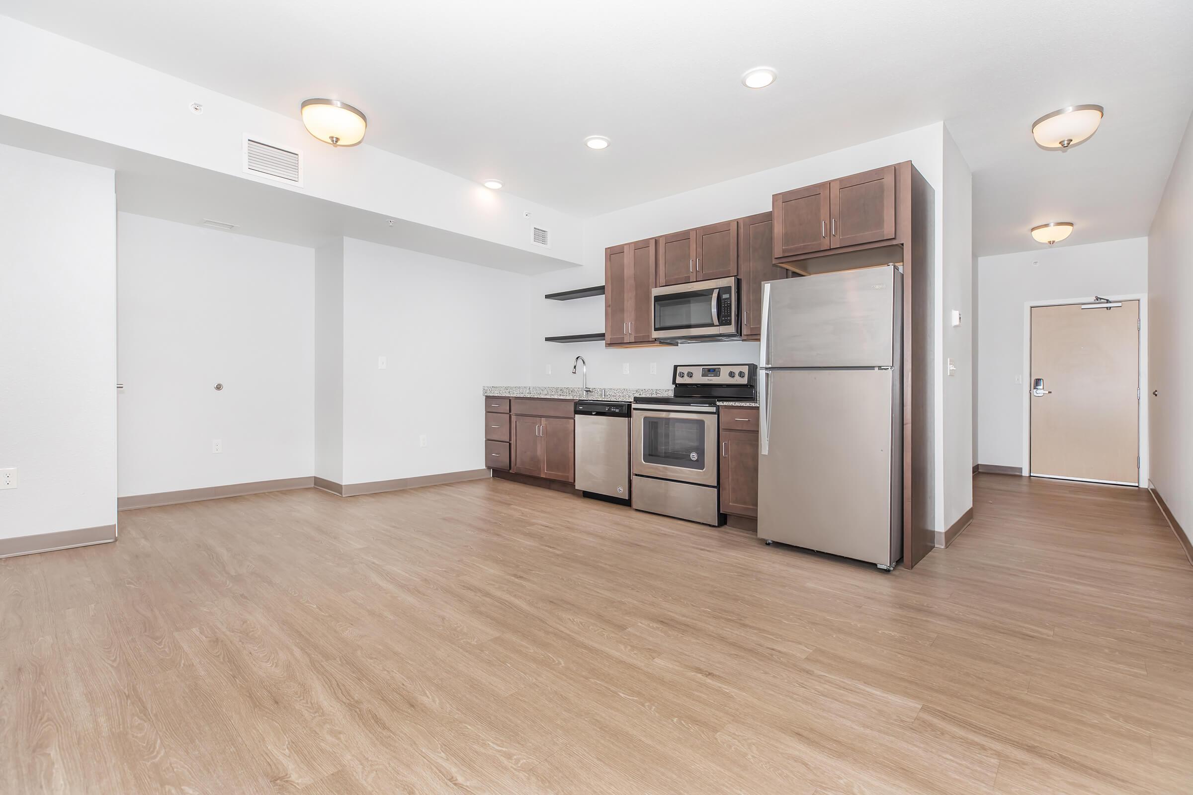 A modern kitchen area featuring stainless steel appliances, including a refrigerator, stove, and microwave, with dark wood cabinetry. The open layout showcases light-colored walls and a spacious floor design. There is a front door visible in the background, leading to another room or hallway.