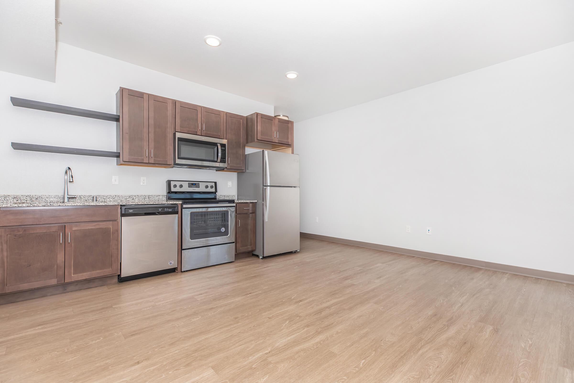 Modern kitchen with dark wood cabinets, stainless steel appliances, and a granite countertop. The space features a stove, microwave, dishwasher, and refrigerator. Light-colored walls and a wooden floor create a bright and open atmosphere.