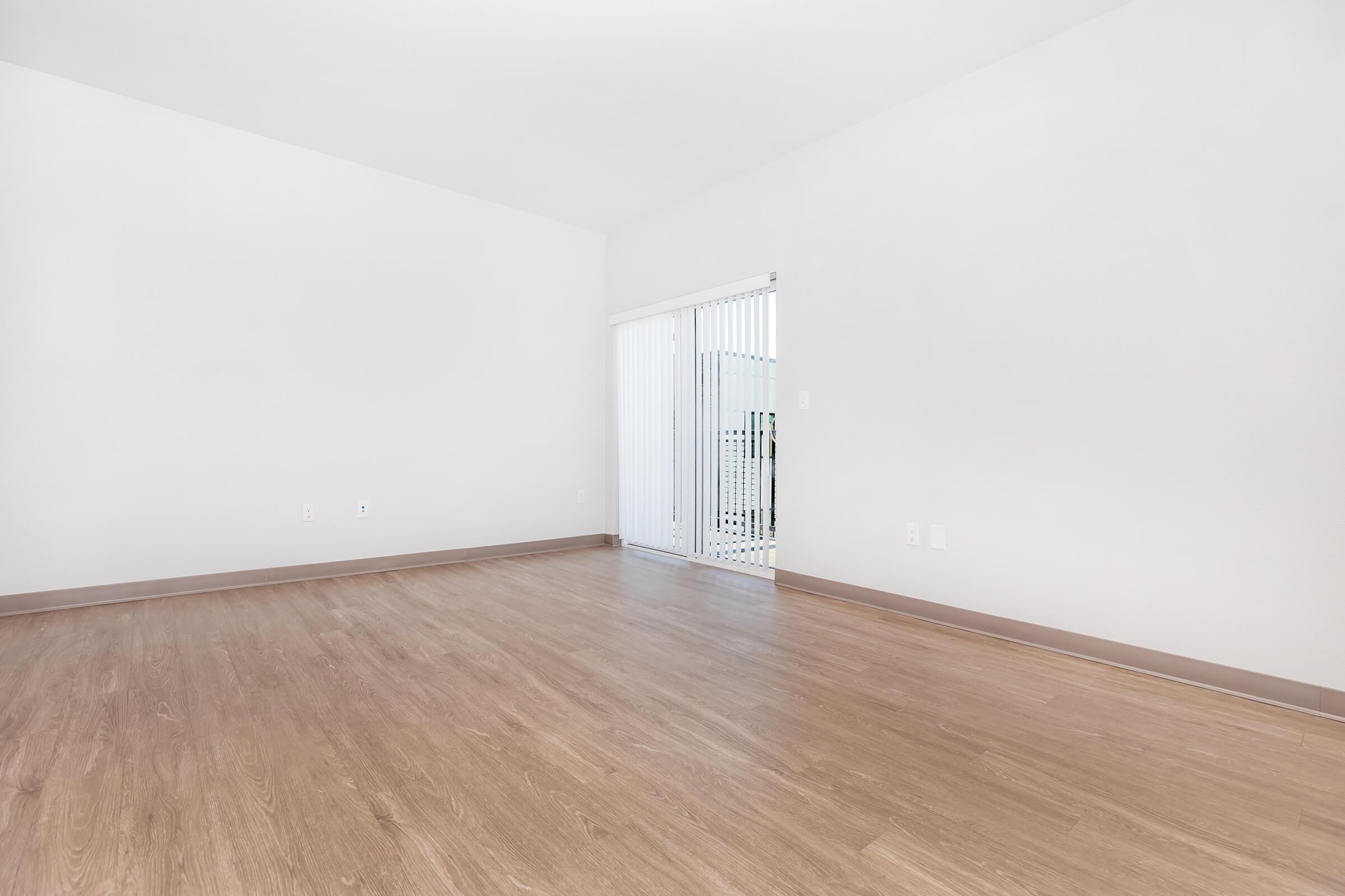A spacious interior room with light-colored walls and a smooth wooden floor. There is a sliding glass door that leads to an outdoor area, allowing natural light to enter the space. The room is empty and has a minimalist design, creating a bright and open atmosphere.