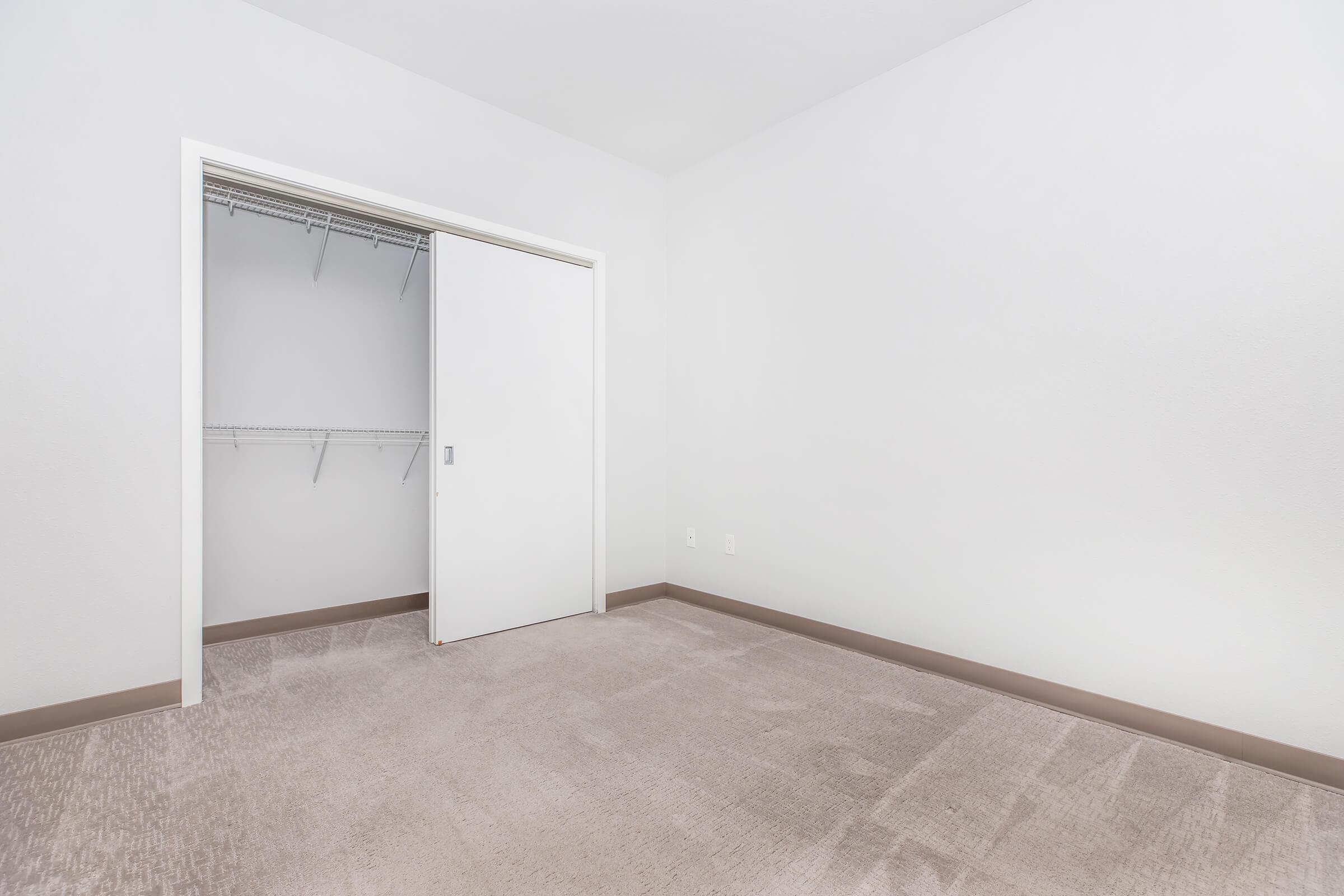 A minimalist room featuring light-colored walls and a beige carpet. There is a spacious, empty closet with sliding white doors to the left. The room has no furniture, creating a clean and open atmosphere, ideal for personal customization or decoration.