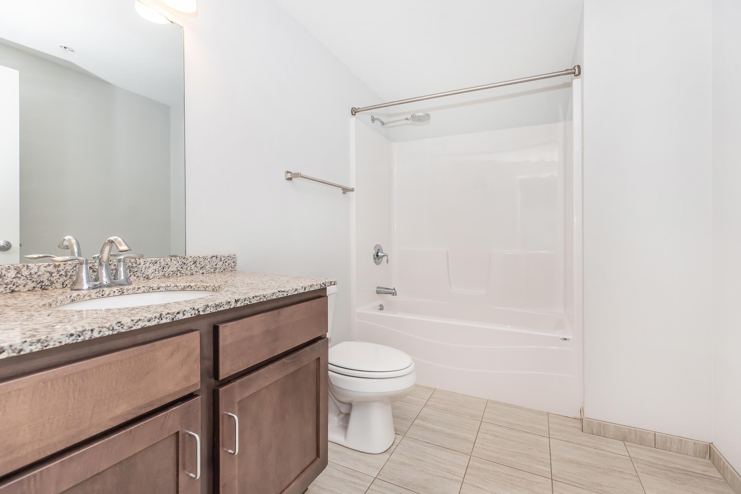 A bathroom featuring a white bathtub with a shower, a granite countertop sink with two faucets, a modern toilet, and tiled flooring. The walls are painted in a light color, and there is a towel bar mounted on the wall. The overall design is clean and contemporary.