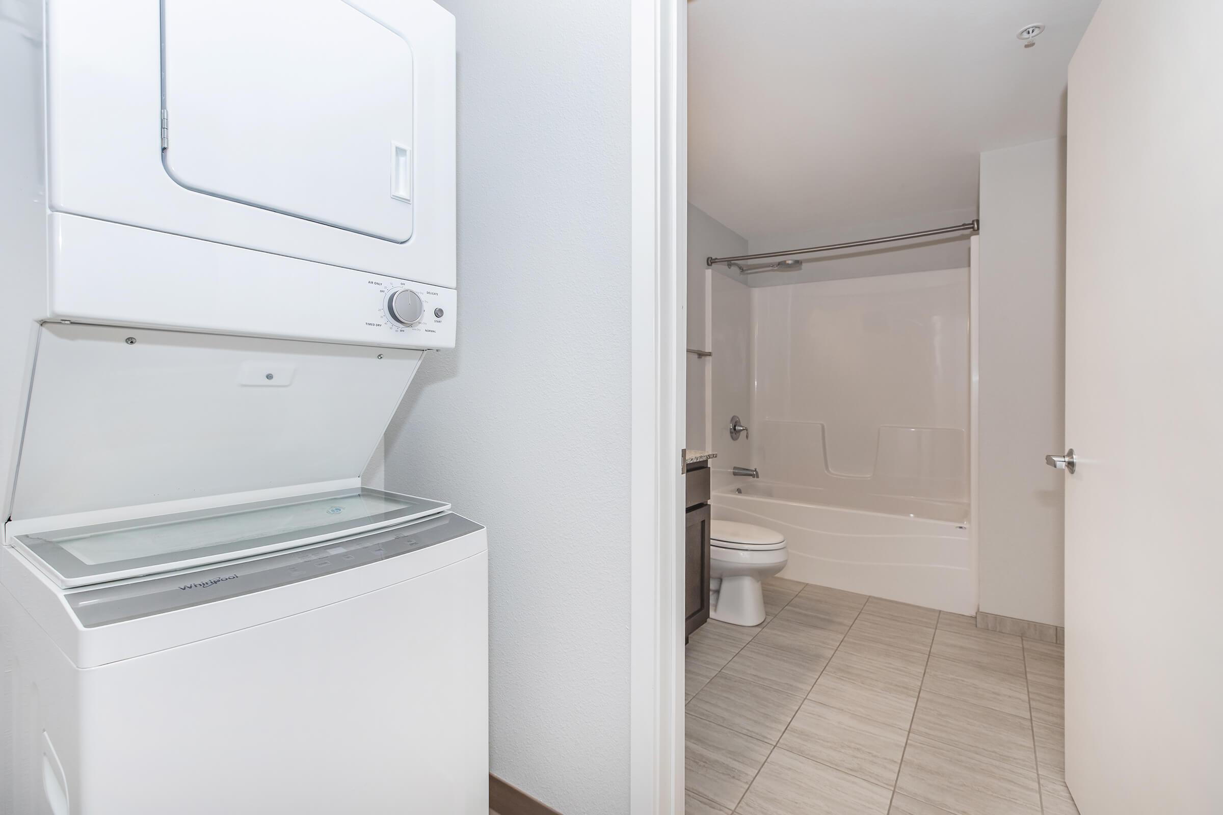 A compact laundry area featuring a stacked washer and dryer to the left, with a bathroom visible on the right. The bathroom includes a shower/tub combo, toilet, and neutral-colored tile flooring. The walls are painted in light tones, creating a clean and modern appearance.