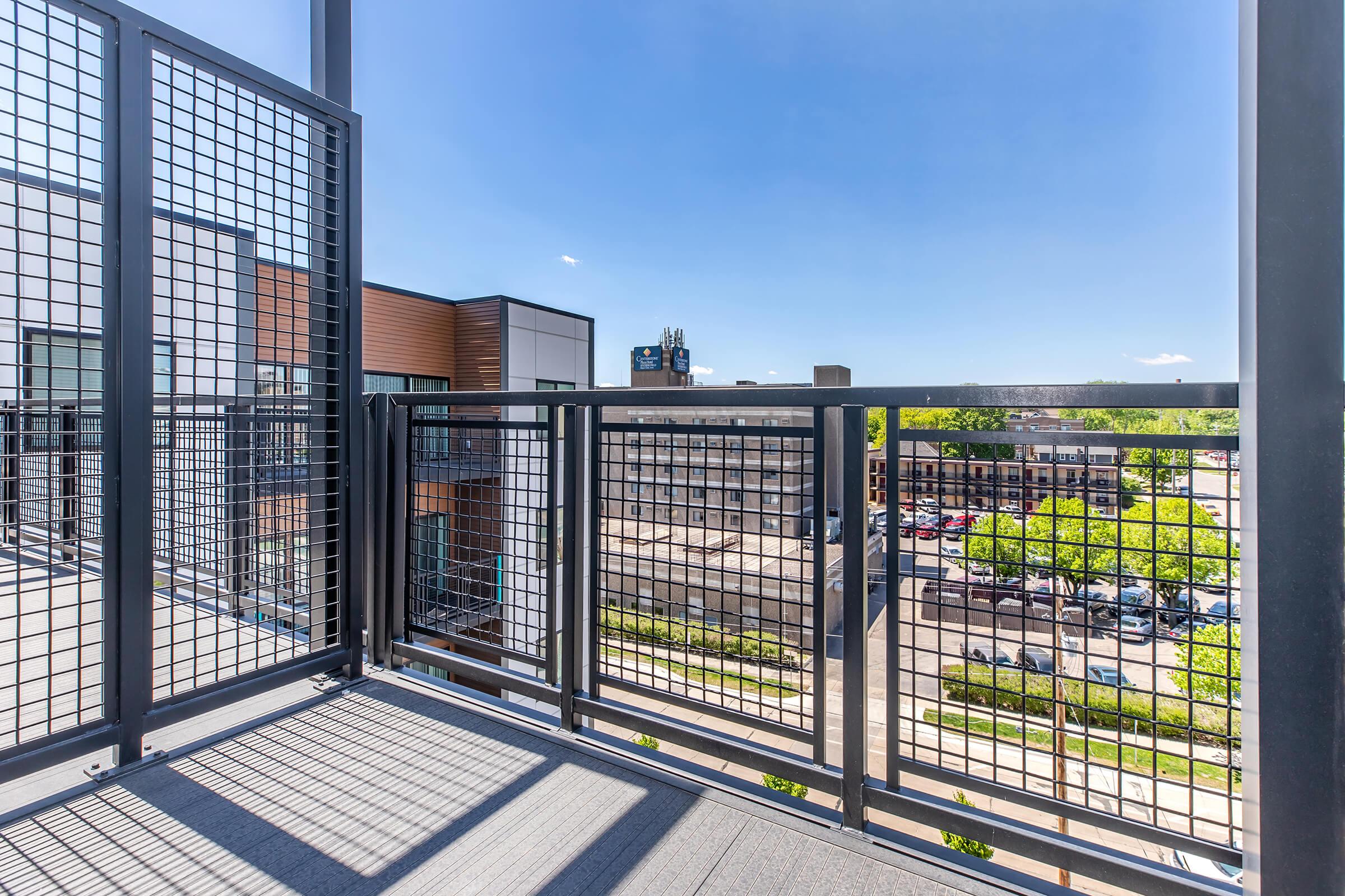 A modern balcony with a metal railing, overlooking a cityscape featuring buildings and parked cars. The sky is clear and blue, with a few clouds visible. Green trees are seen in the foreground, adding a touch of nature to the urban environment.