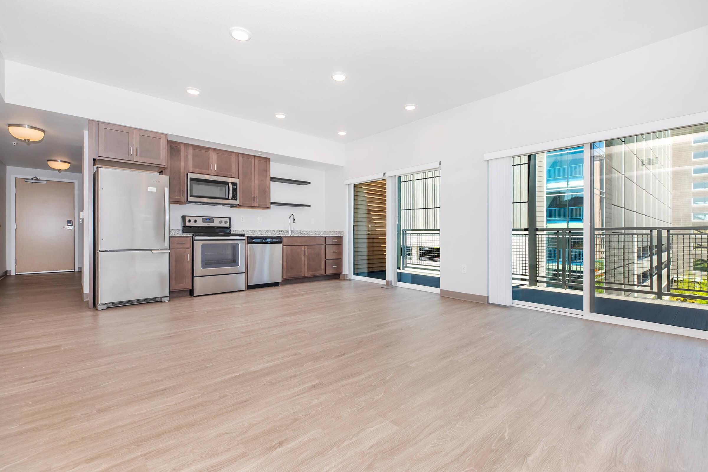 Bright and spacious modern apartment interior featuring an open-plan kitchen with stainless steel appliances, wooden cabinetry, and a large living area with wood-style flooring. Natural light streams in through sliding glass doors that lead to a balcony, providing a view of the building's exterior.