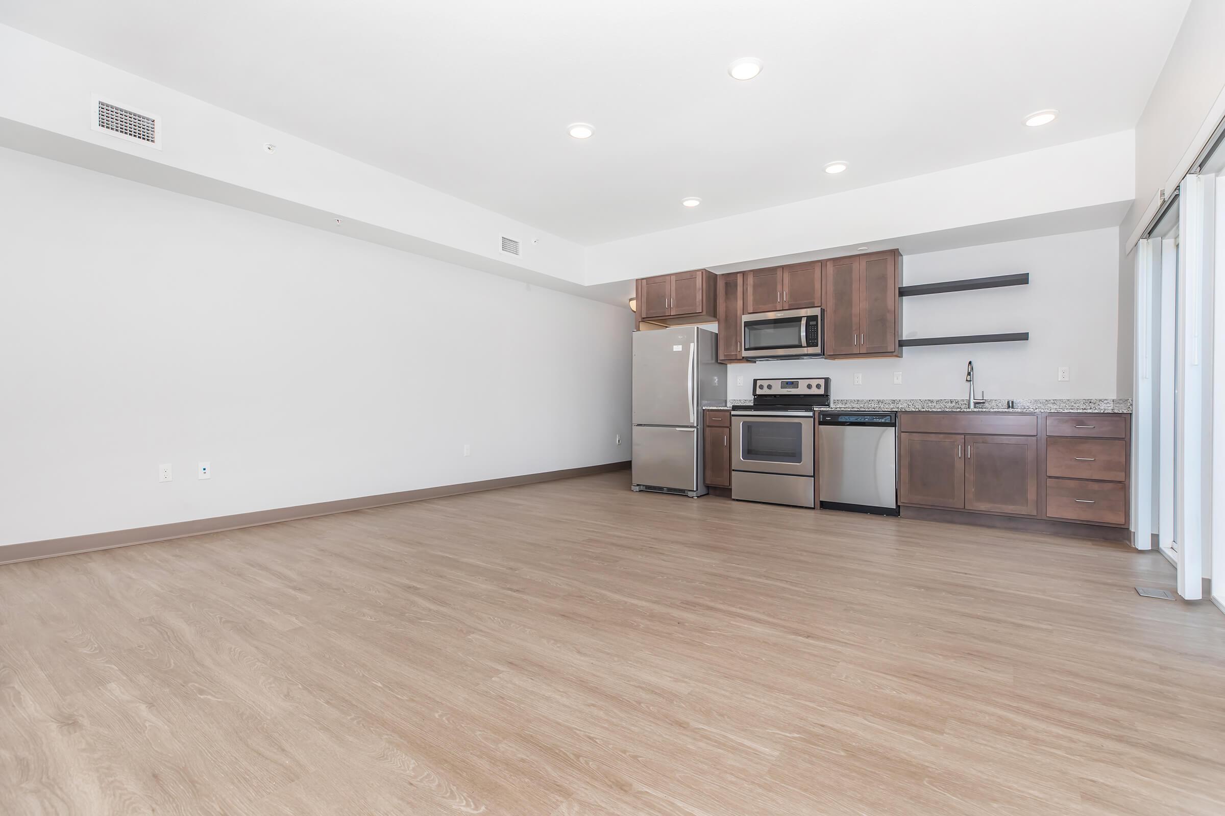 Spacious modern apartment interior featuring a kitchen with stainless steel appliances, including a refrigerator, oven, and microwave. Light wood flooring throughout, neutral walls, and large windows allowing natural light. Open floor plan with ample space for furniture arrangement.