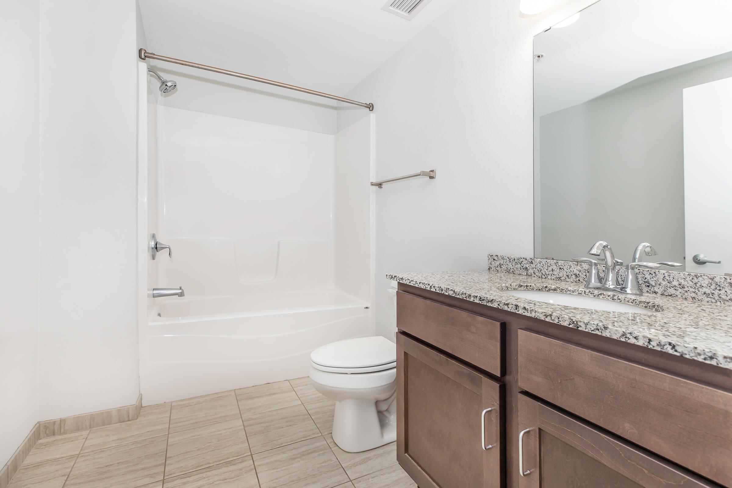 Modern bathroom featuring a beige tiled floor, a shower-tub combo with a white surround, and a granite countertop vanity. The bathroom has a large mirror above the sink and light fixtures mounted on the ceiling, creating a bright and spacious atmosphere.
