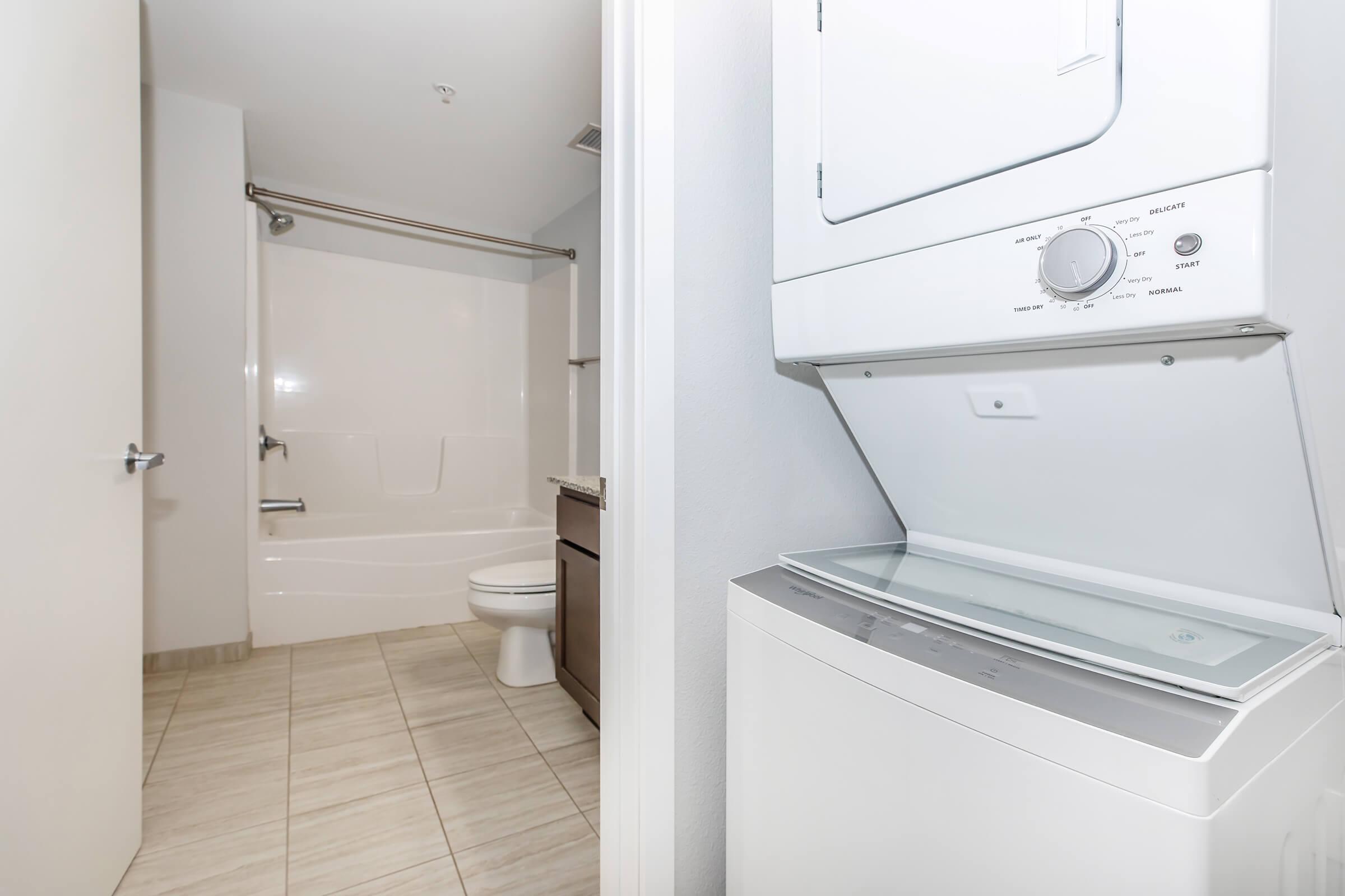 A view of a modern bathroom featuring a bathtub and a toilet, with a laundry unit stacked above a washer below. The space has light-colored tiles and neutral walls, creating an open and clean atmosphere. There is a door leading out, enhancing the room's accessibility.