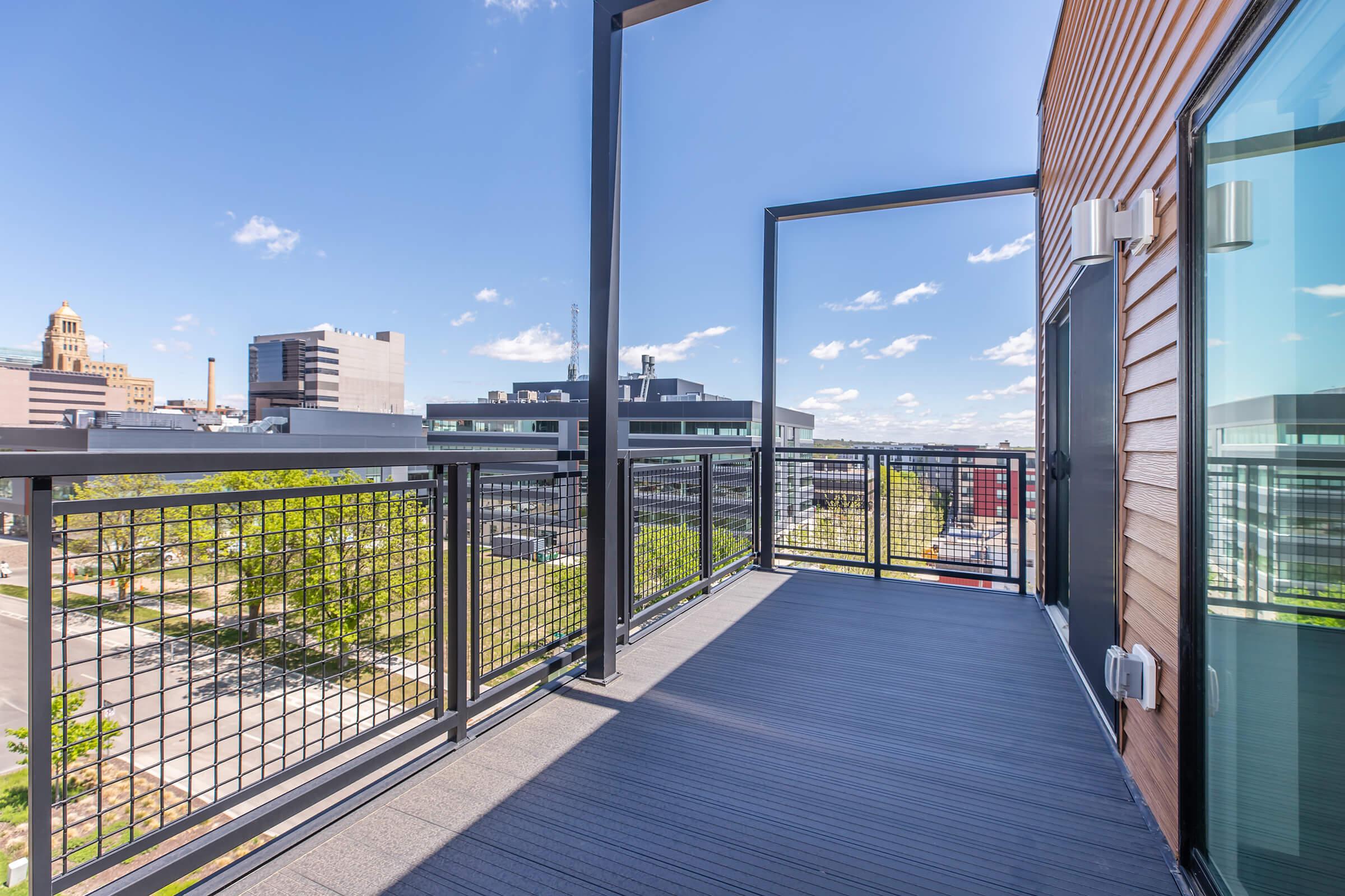 A modern balcony with a black metal railing overlooks a cityscape featuring buildings and blue sky with scattered clouds. The balcony has a sleek floor and a glass wall, creating a spacious and inviting outdoor area.