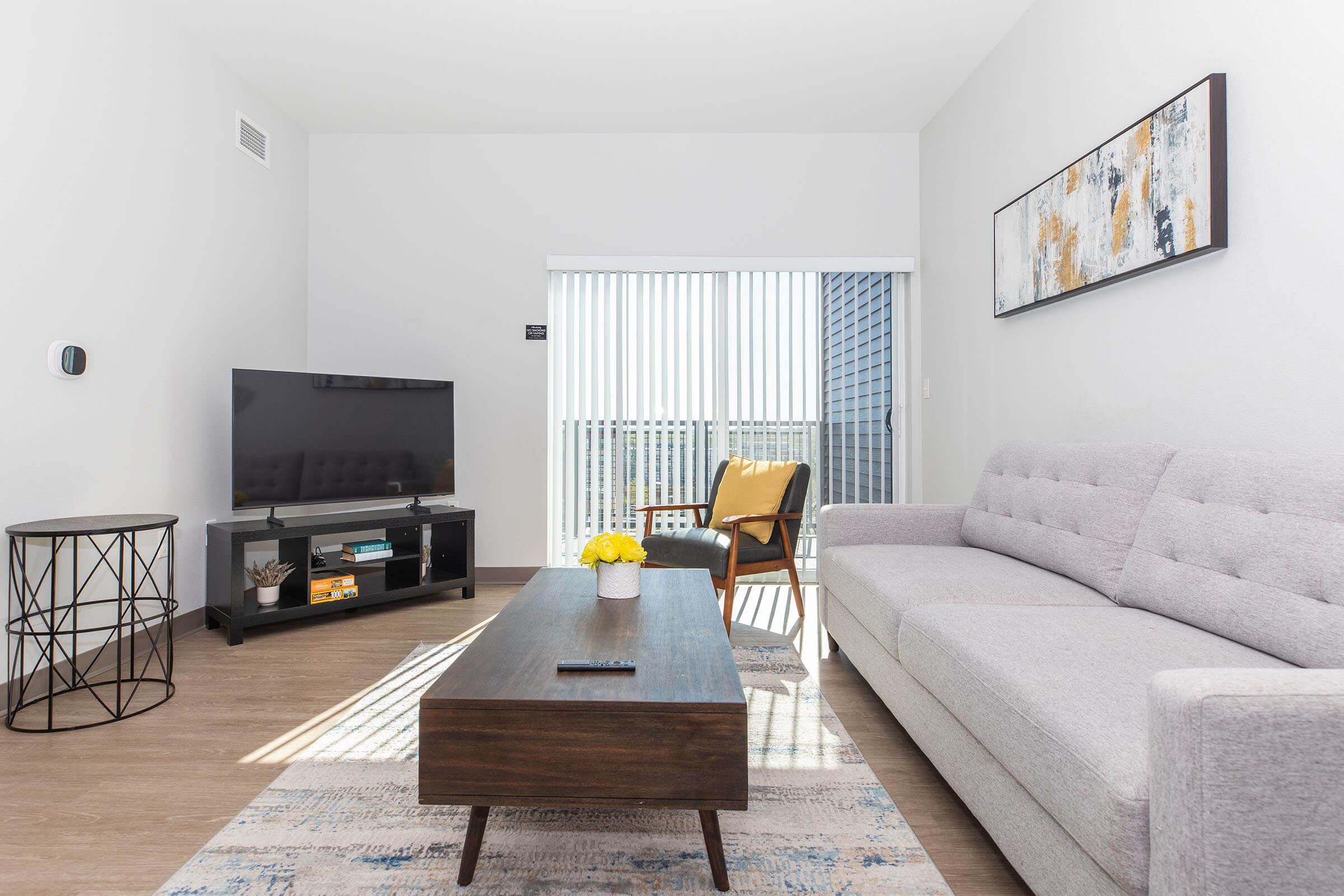 Modern living room featuring a gray sofa, a wooden coffee table, and a stylish chair. A flat-screen TV is on a black media stand, and a decorative table sits nearby. Large sliding glass doors lead to a balcony, allowing natural light to brighten the space. Wall art adds a touch of color to the minimalist decor.