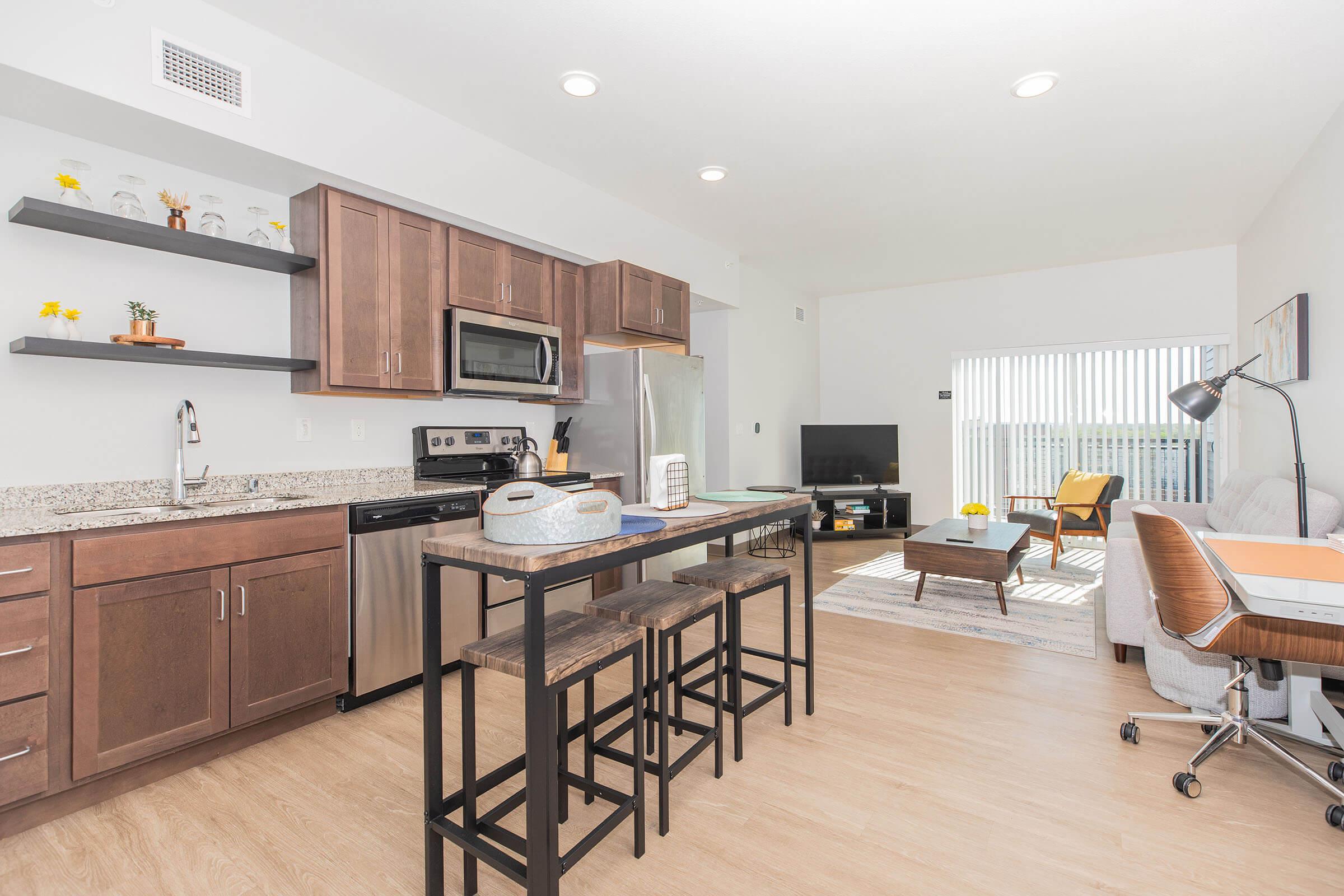 Modern kitchen and living area featuring wooden cabinetry, stainless steel appliances, and a granite countertop bar with stools. The room is bright and airy, with a large window allowing natural light, a TV in the corner, and a comfortable seating area with a chair and a desk.