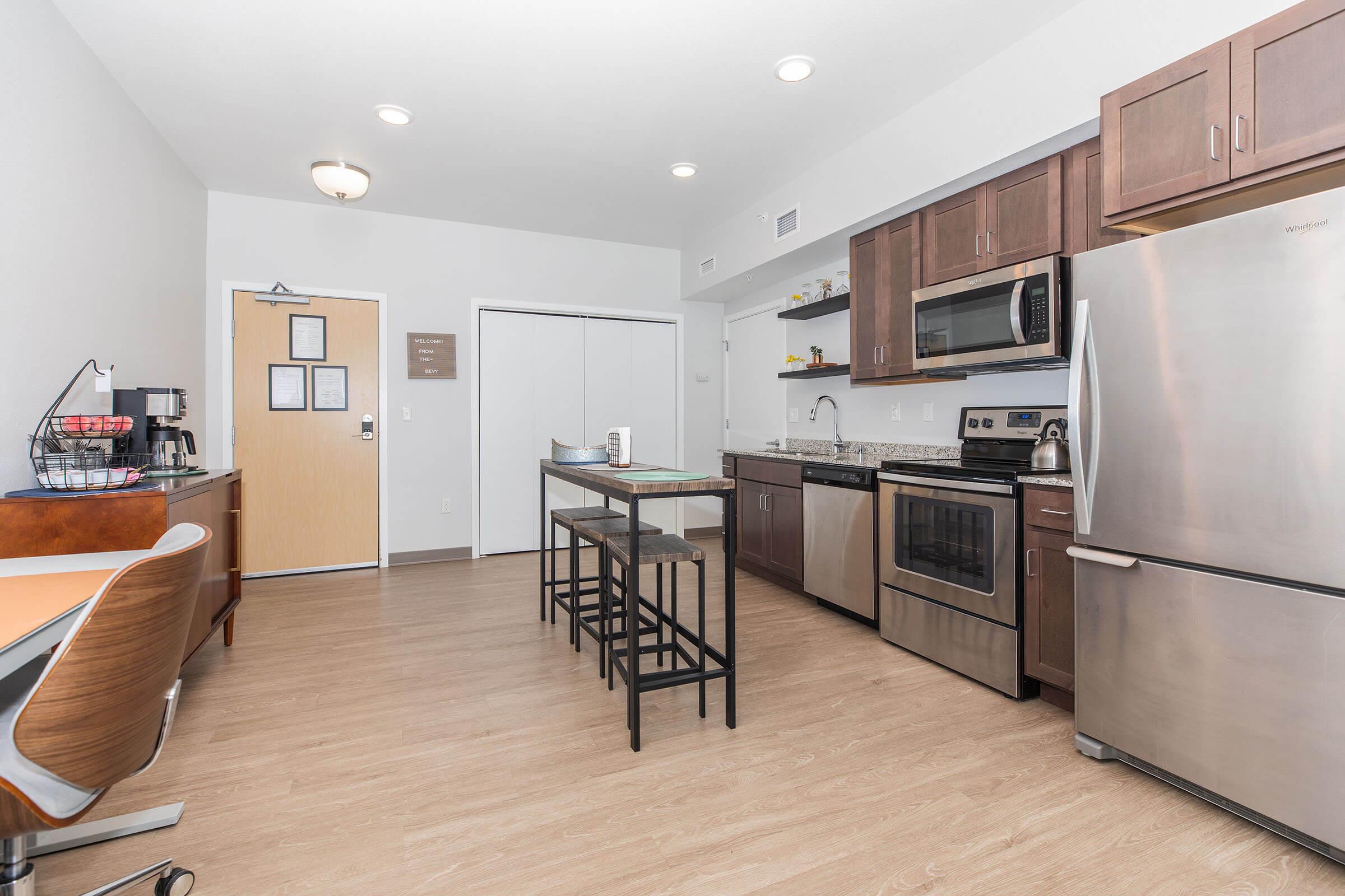 A modern kitchen featuring stainless steel appliances, including a refrigerator, oven, and microwave. There's a granite countertop with bar seating for four, a small desk area, and light wooden flooring. The entry door is visible, along with a closet and light fixtures overhead.