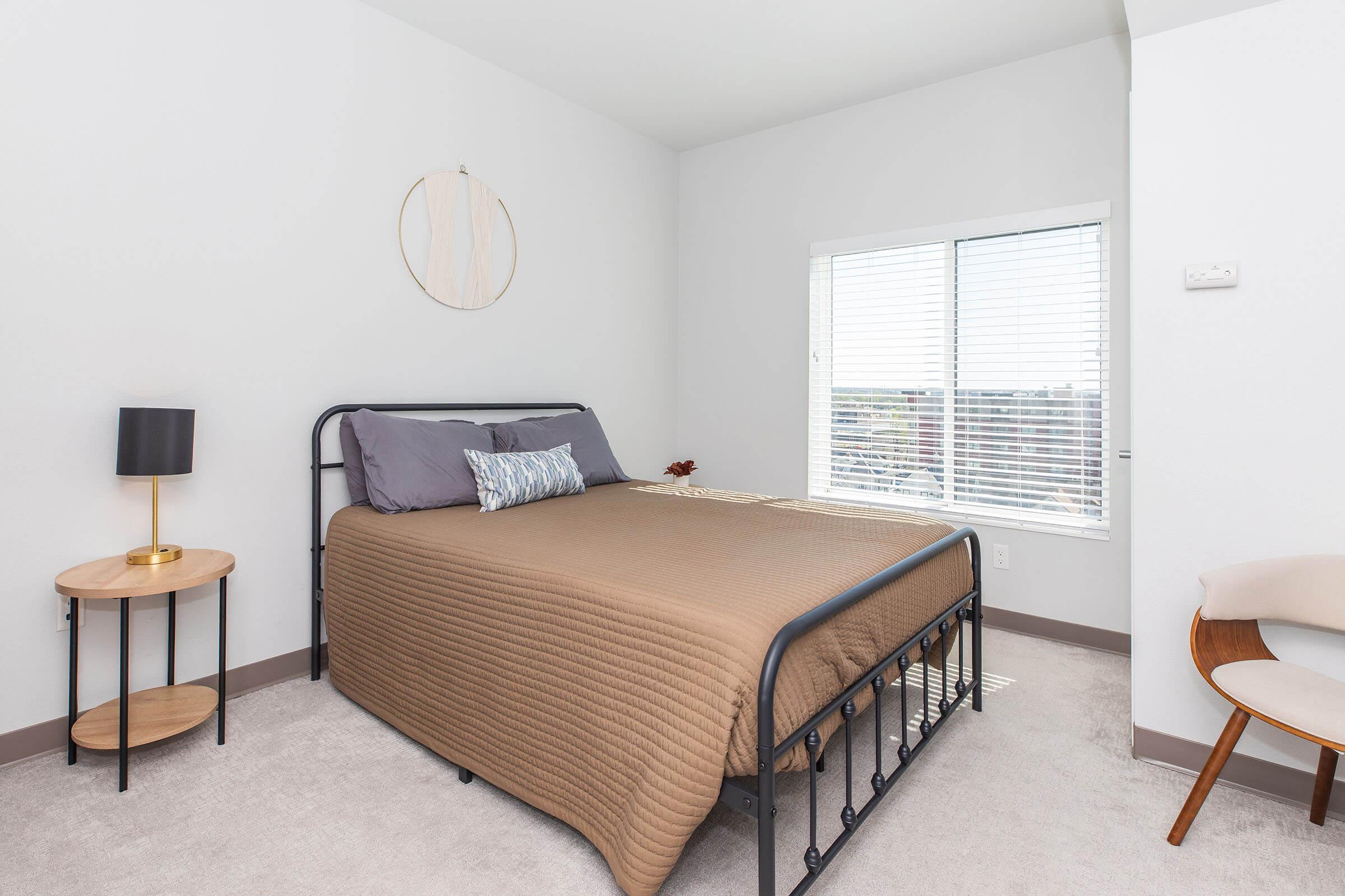 A modern bedroom featuring a black metal bed frame with a brown quilt, purple and blue accent pillows, and a side table with a black lamp. A large window with blinds allows natural light to enter, while a round wall decor piece hangs above the bed. The room has light-colored walls and carpet.