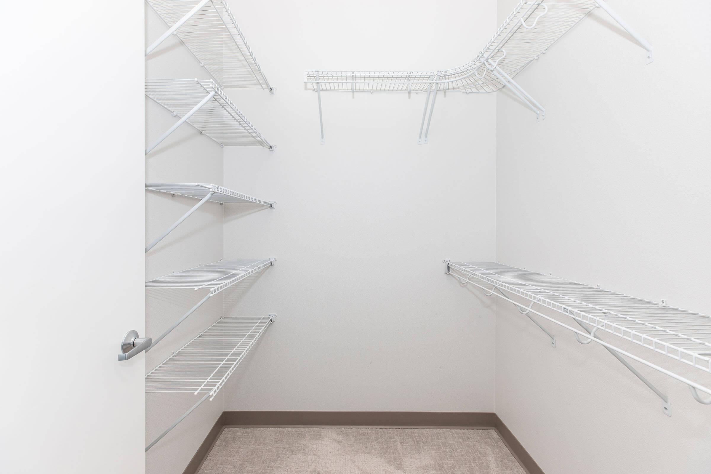 A spacious empty closet with wire shelving along the walls, featuring multiple shelves and a neutral-colored carpet floor. The walls are painted white, and the closet appears well-lit and tidy, offering ample storage space.