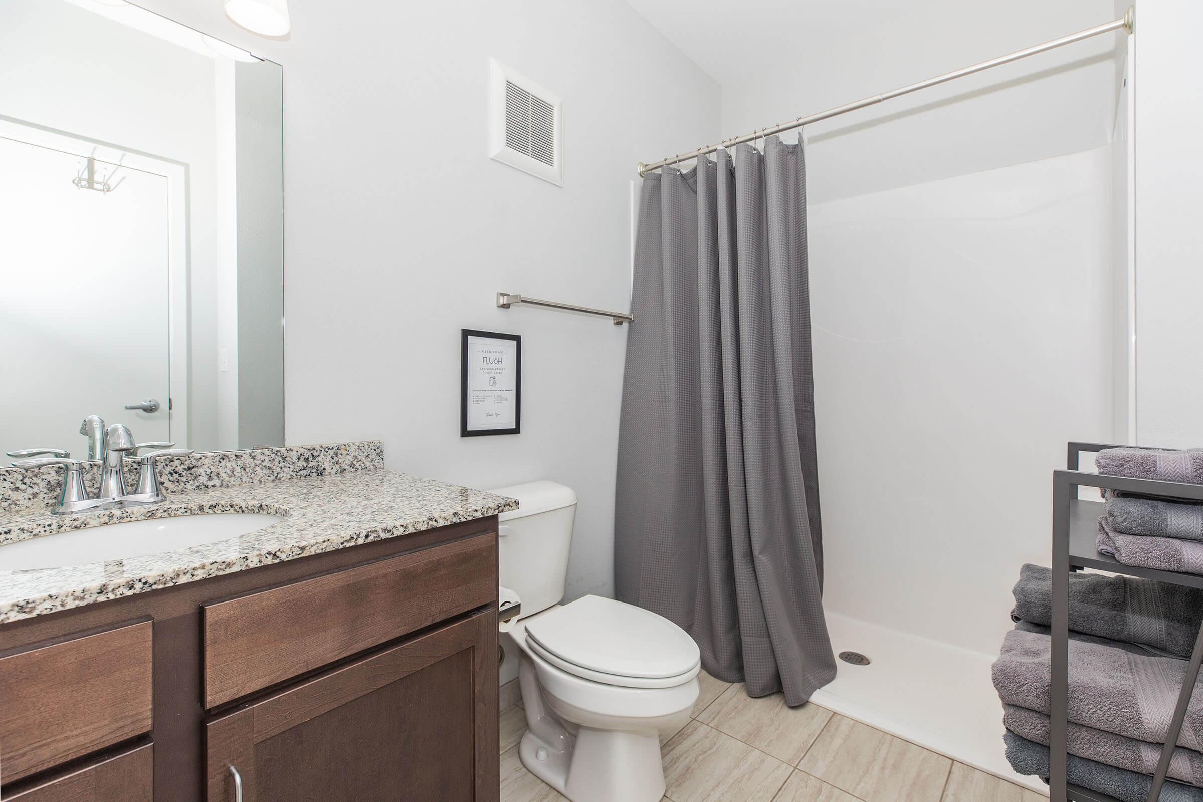 A modern bathroom featuring a granite countertop sink, white toilet, and a shower with a gray curtain. There is a stack of neatly arranged gray towels and a wall-mounted sheet with information. The flooring is tiled in a light color, and the walls are painted white for a clean, bright look.