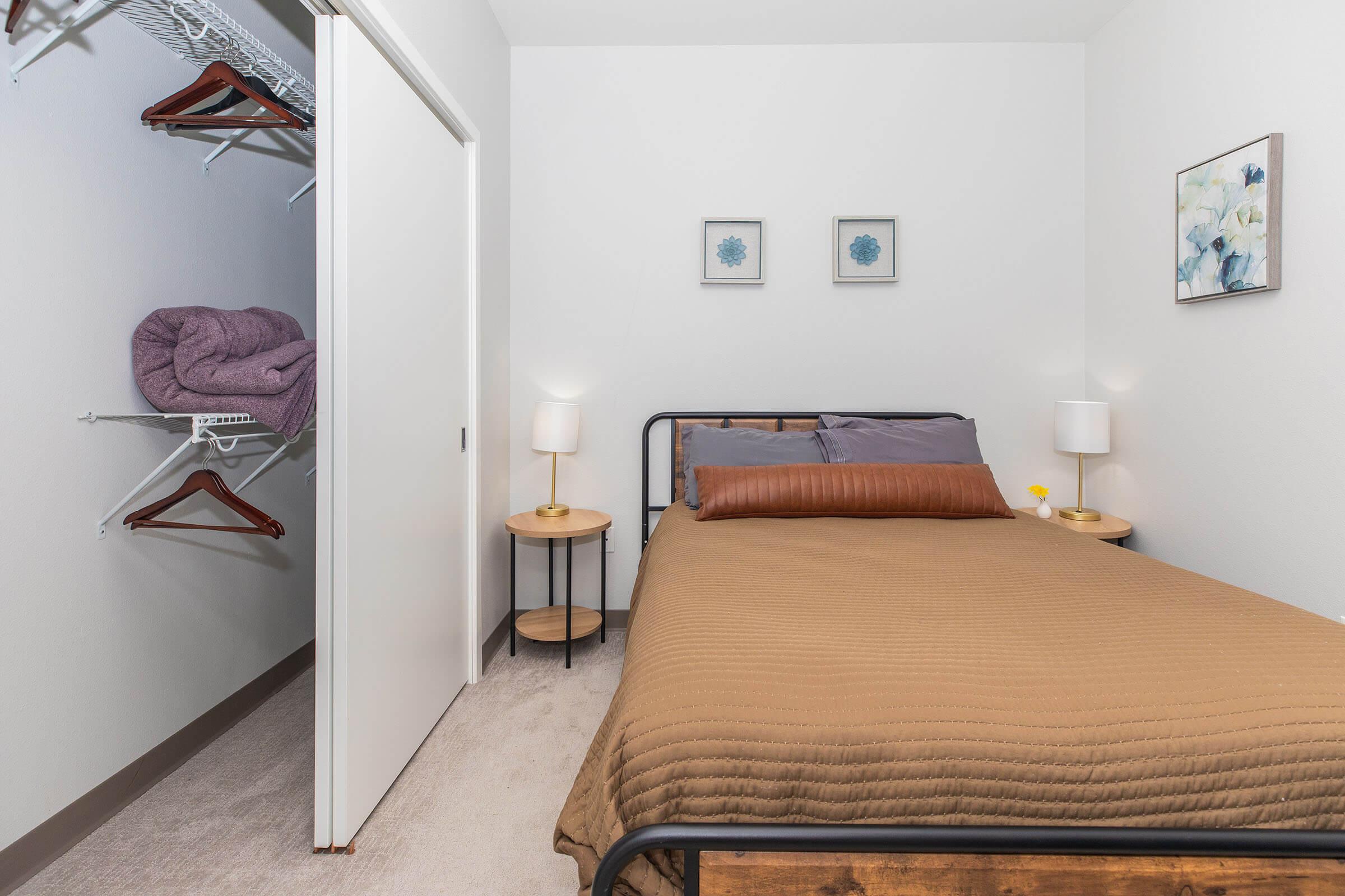 Cozy bedroom featuring a bed with brown bedding, two bedside tables with lamps, and wall art. A closet is visible on the left with hangers and folded blankets. The room has neutral-colored walls and carpet, providing a simple and inviting atmosphere.