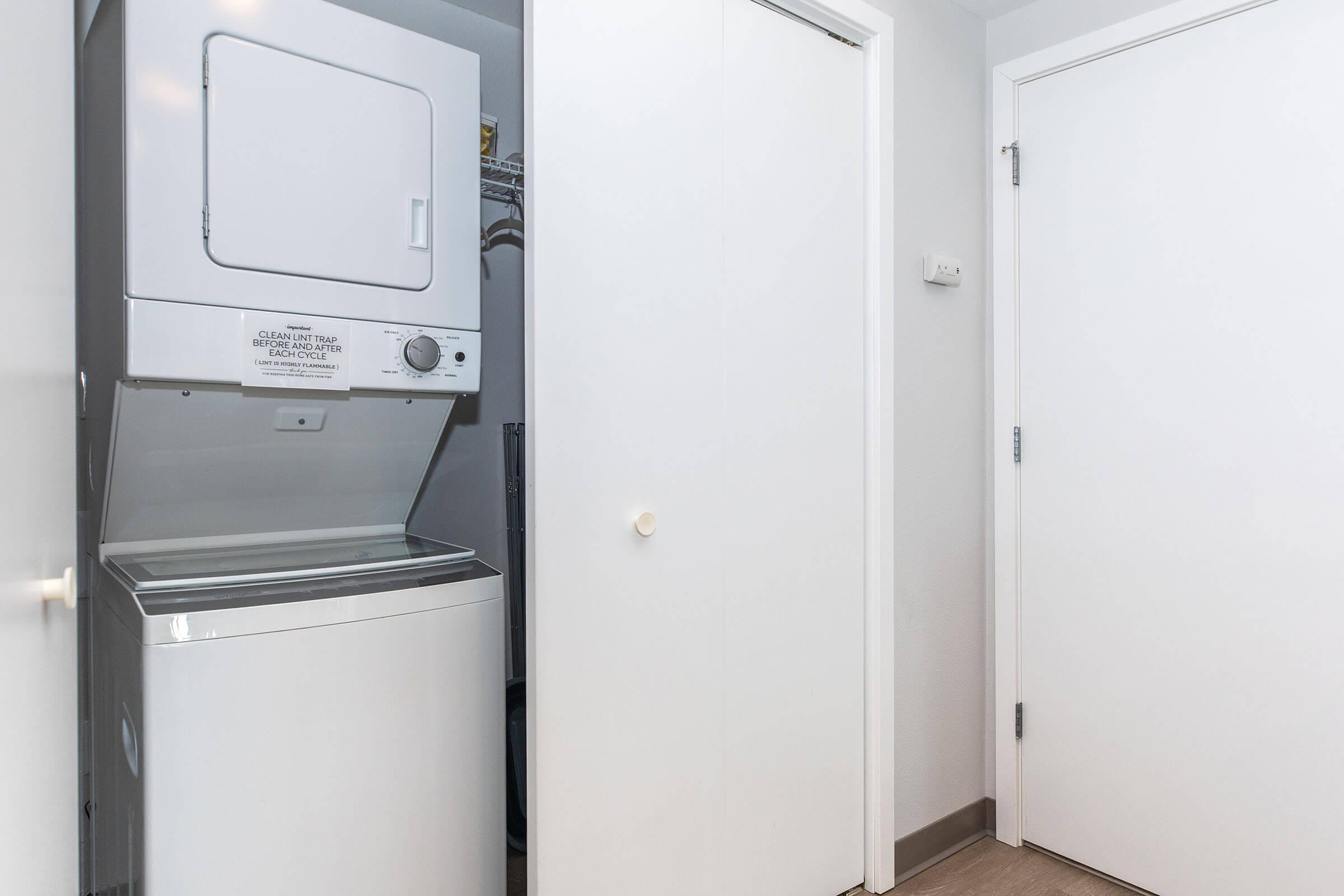 A stacked washer and dryer unit in a small laundry area with white walls and a closed wooden door. The floor is light-colored, and the space appears clean and organized.