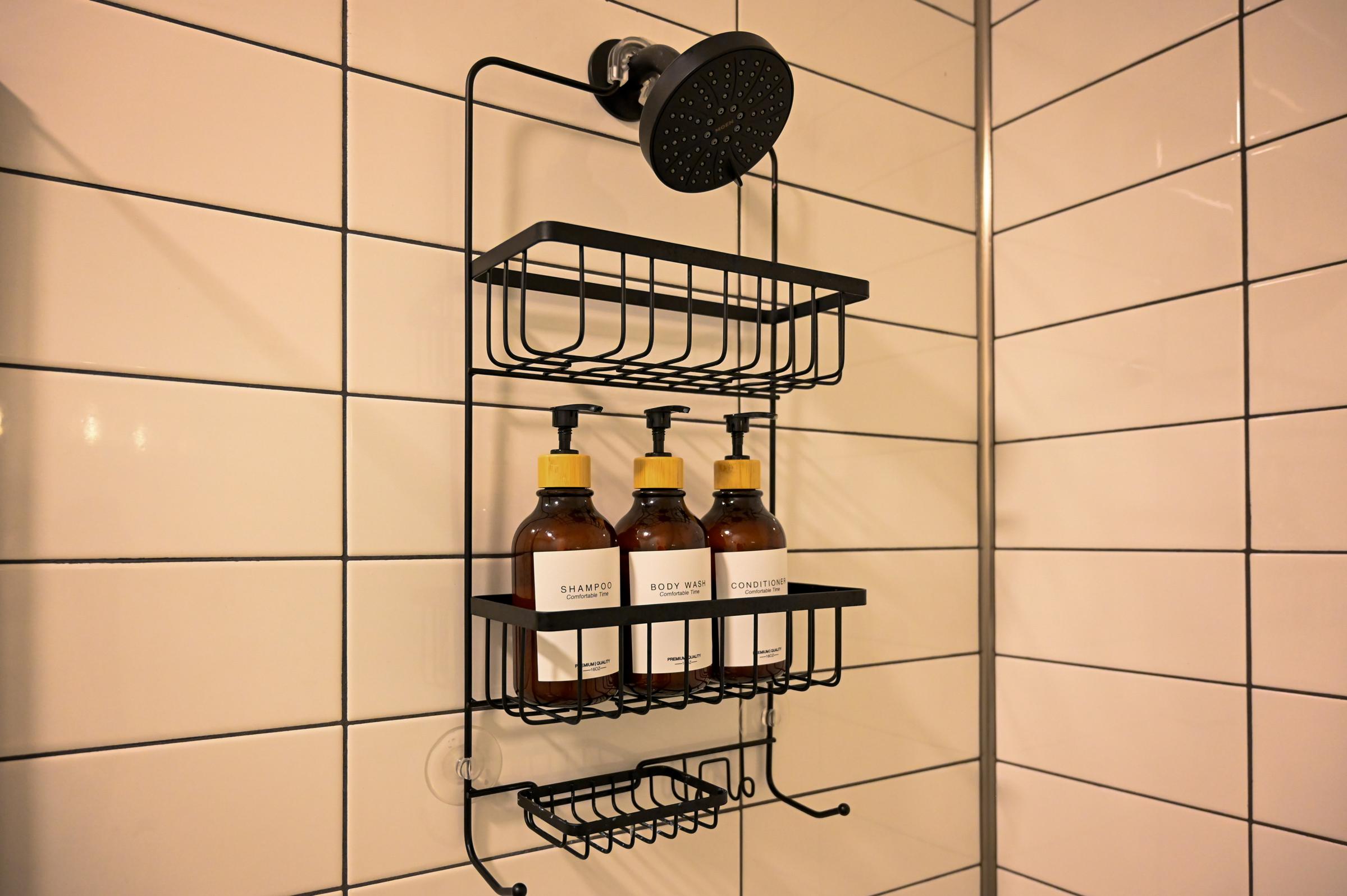 A sleek black shower caddy mounted on white tiled walls, featuring three amber-colored bottles labeled "Shampoo," "Body Wash," and "Conditioner." Above the caddy, a modern shower head is visible, creating a contemporary bathroom aesthetic.