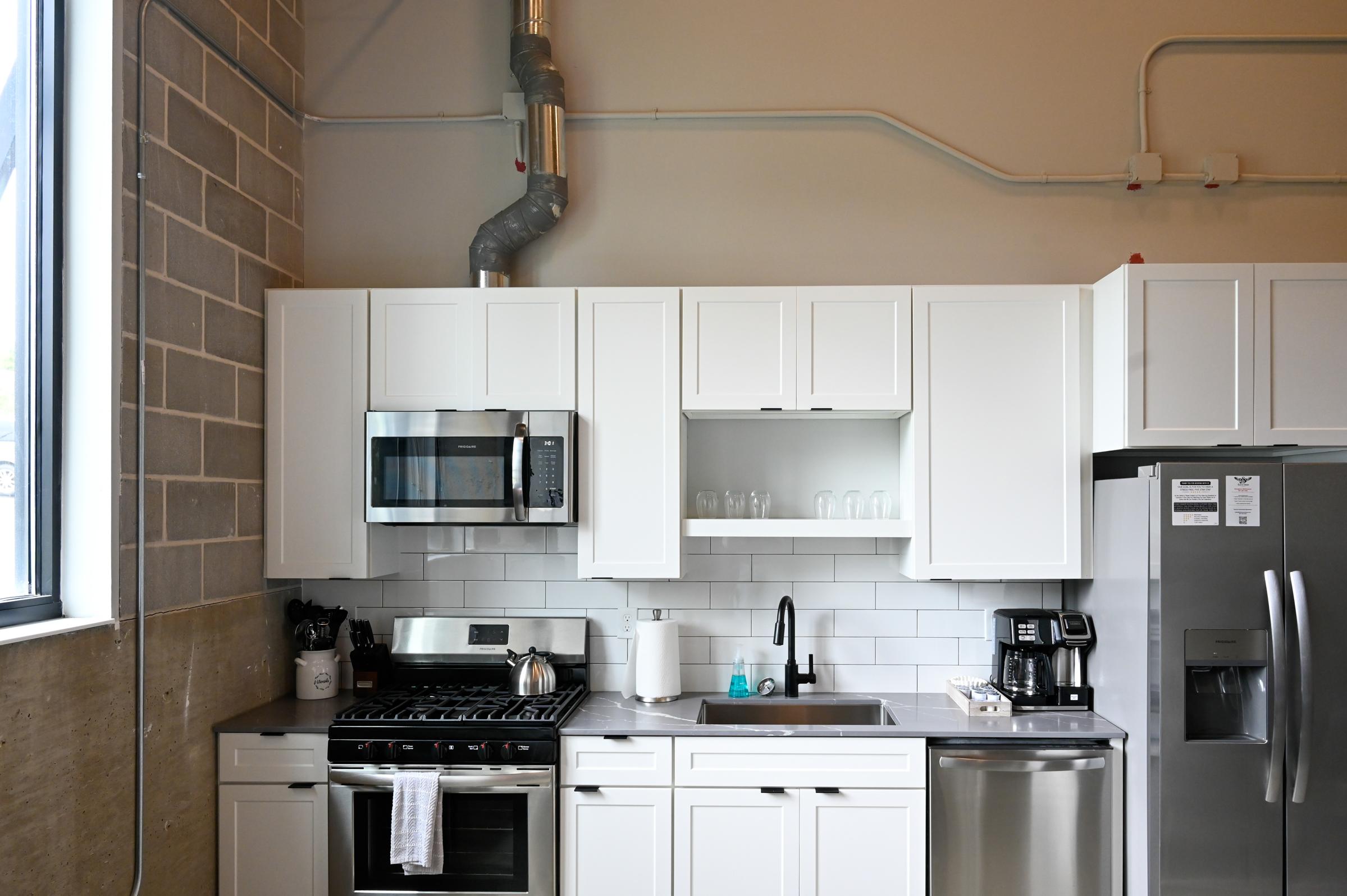 A modern kitchen featuring white cabinets, a stainless steel microwave, an oven, and a dishwasher. The countertop is equipped with a sink and a coffee maker. Clear glasses are visible on the shelf above the sink. The kitchen has a minimalist design with exposed pipes and a mix of light and neutral tones.