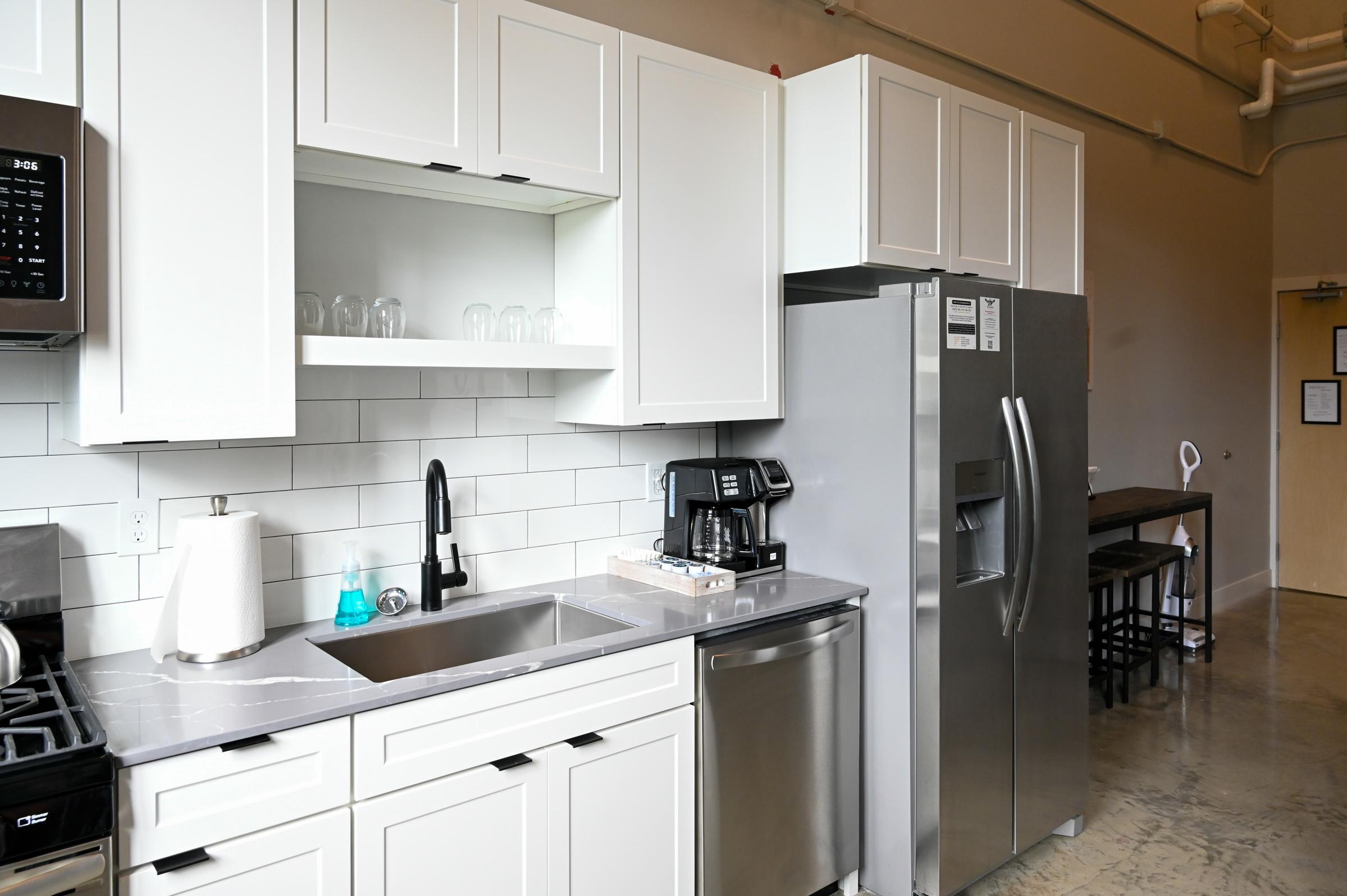 Modern kitchen featuring white cabinetry, a stainless steel refrigerator, and a sleek countertop with a sink. The space includes a coffee maker and a glass dishware display. The flooring is polished concrete, and there is a dining area with a wooden table and chairs in the background.