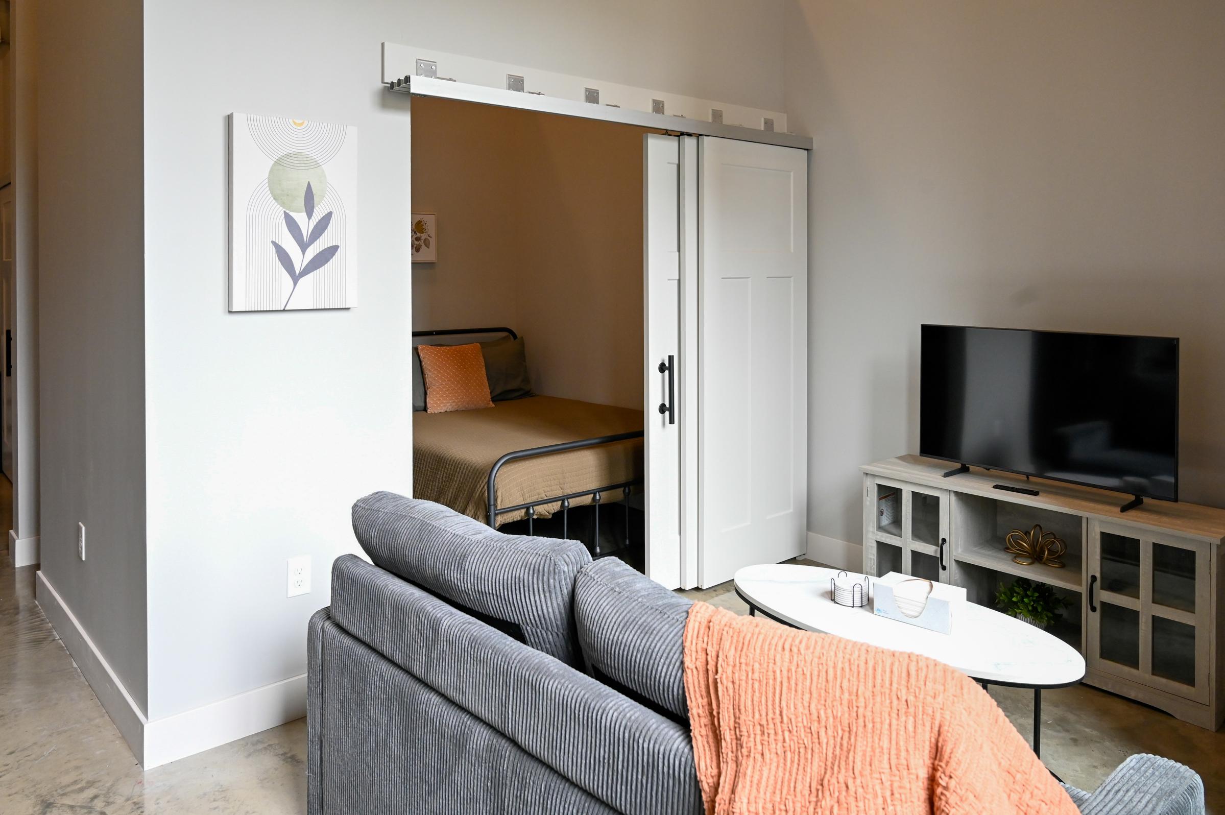 A cozy interior showing a living area with a gray sofa, a coffee table, and a television. In the background, there is a sliding door leading to a bedroom with a bed visible. A piece of wall art hangs on the light gray wall, and a soft orange throw blanket is draped over the sofa. The space is modern and well-lit.