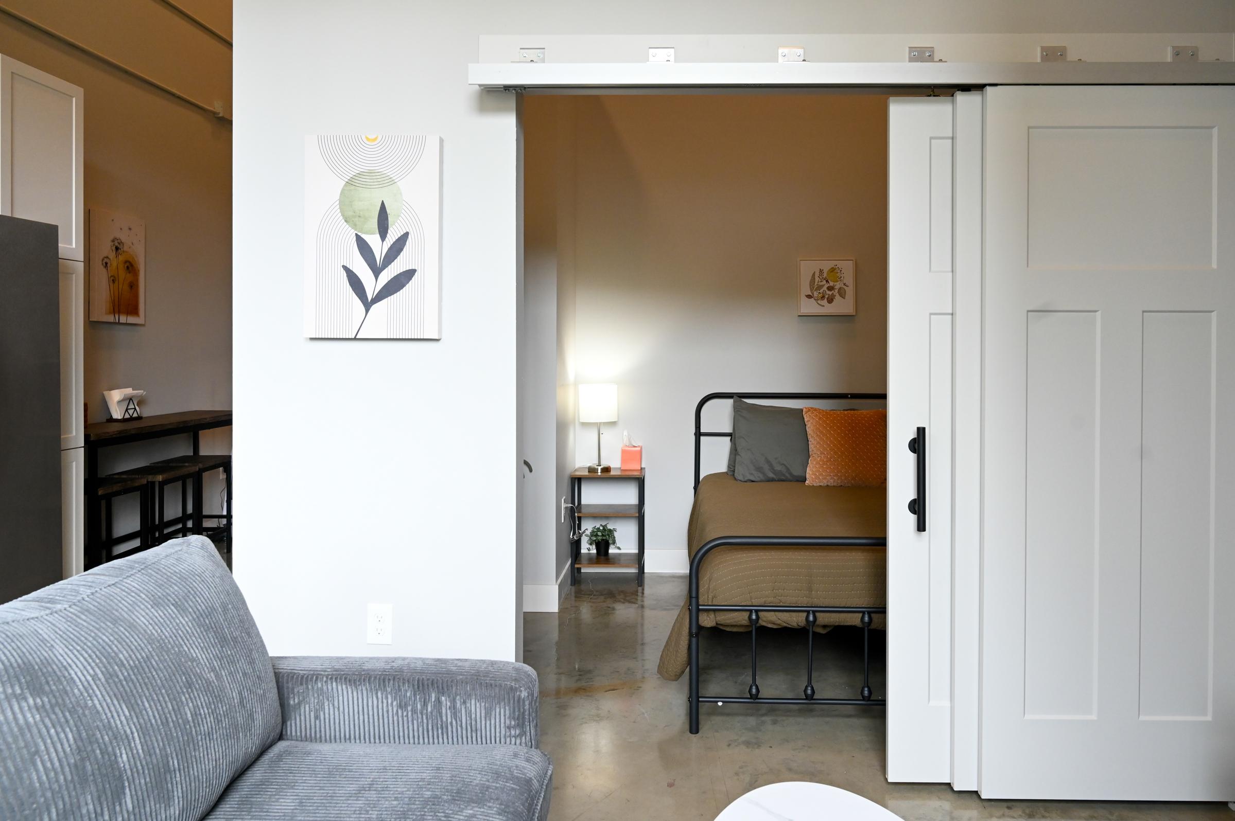 A cozy interior featuring a living area with a gray couch and a small white table. There's a sliding door partially open to reveal a bedroom with a bed, nightstand, and lamp. Artwork hangs on the wall above the couch, and modern decor adds a welcoming atmosphere to the space.