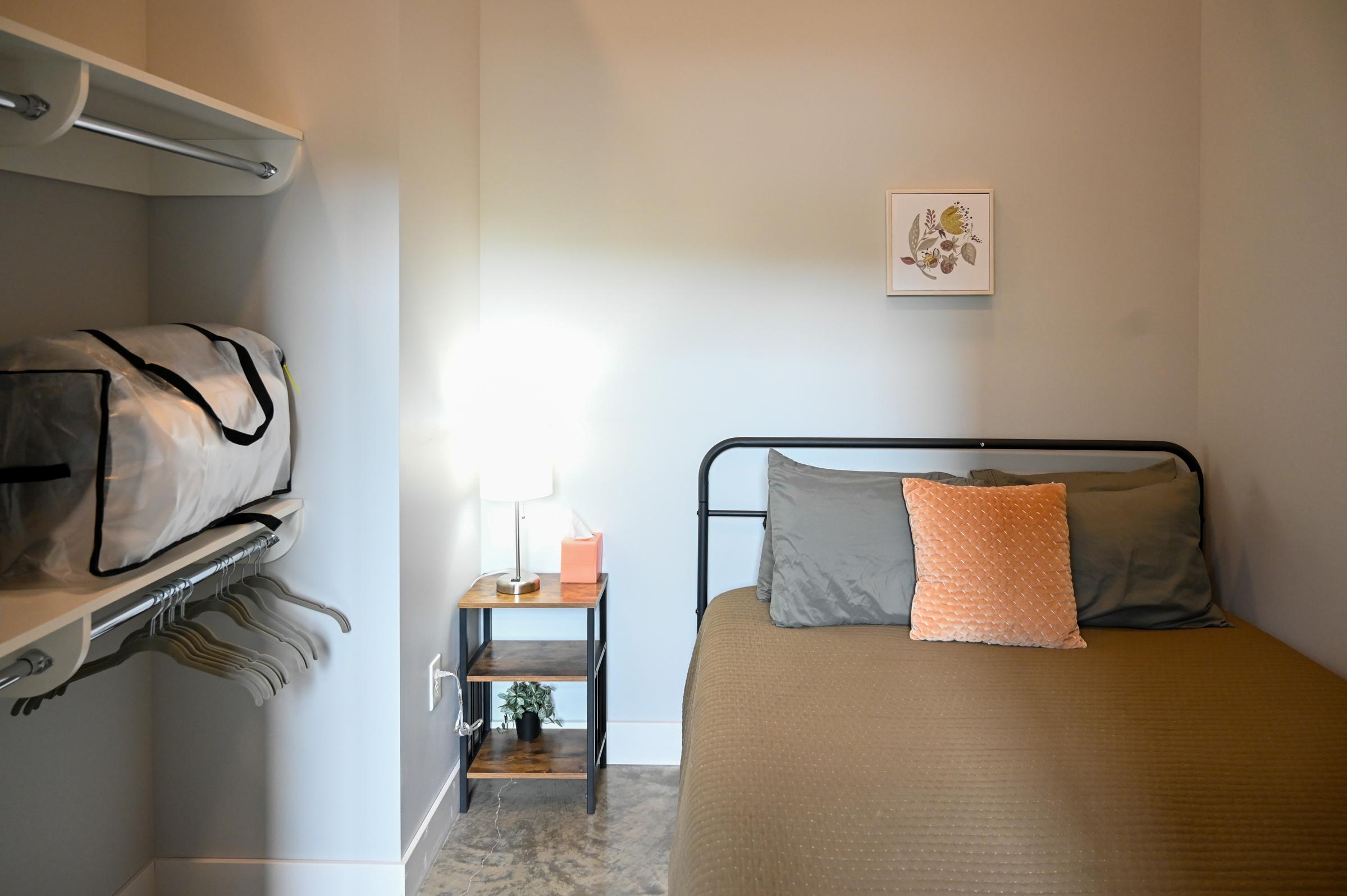A small, elegantly designed bedroom featuring a bed with gray bedding and an orange accent pillow. There’s a side table with a lamp next to the bed, and a wall-mounted shelf with hangers. A framed botanical print is displayed on the wall. The room has neutral-colored walls and a cozy feel.