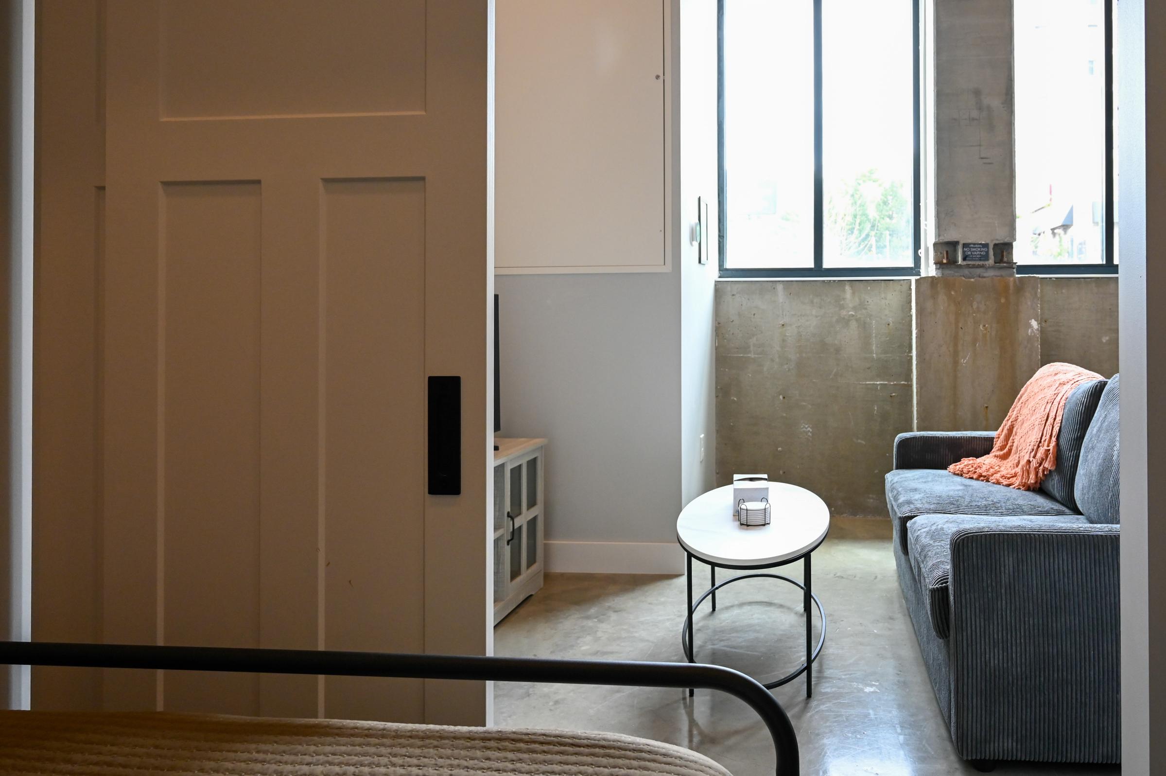 A cozy, modern living space featuring a gray sofa with an orange throw blanket, a round coffee table, and large windows providing natural light. The room has a minimalist aesthetic with concrete walls and a door partially open, revealing the inviting atmosphere.