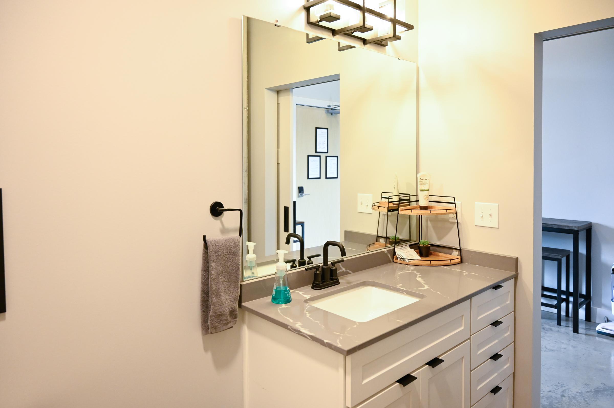 A modern bathroom featuring a gray countertop with a white sink, sleek black faucets, and a towel hanging on a rack. A mirror with stylish lighting above the sink complements the clean design. In the background, a door leads to another room, and a circular shelf displays toiletries.