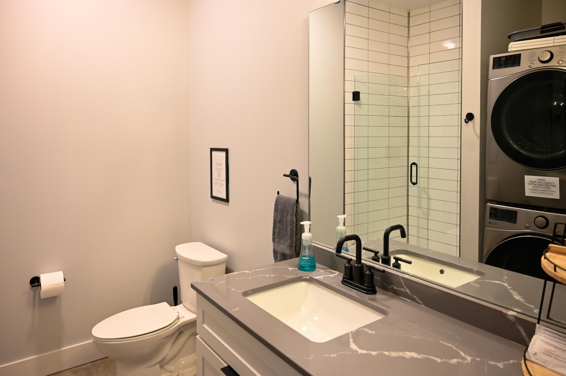 A modern bathroom featuring a white toilet, a sleek sink with a gray countertop, and a large mirror. There is a wall-mounted black faucet and a soap dispenser. To the side, a stacked washer and dryer are visible, and a towel hangs on a black holder. Light gray walls complete the contemporary look.